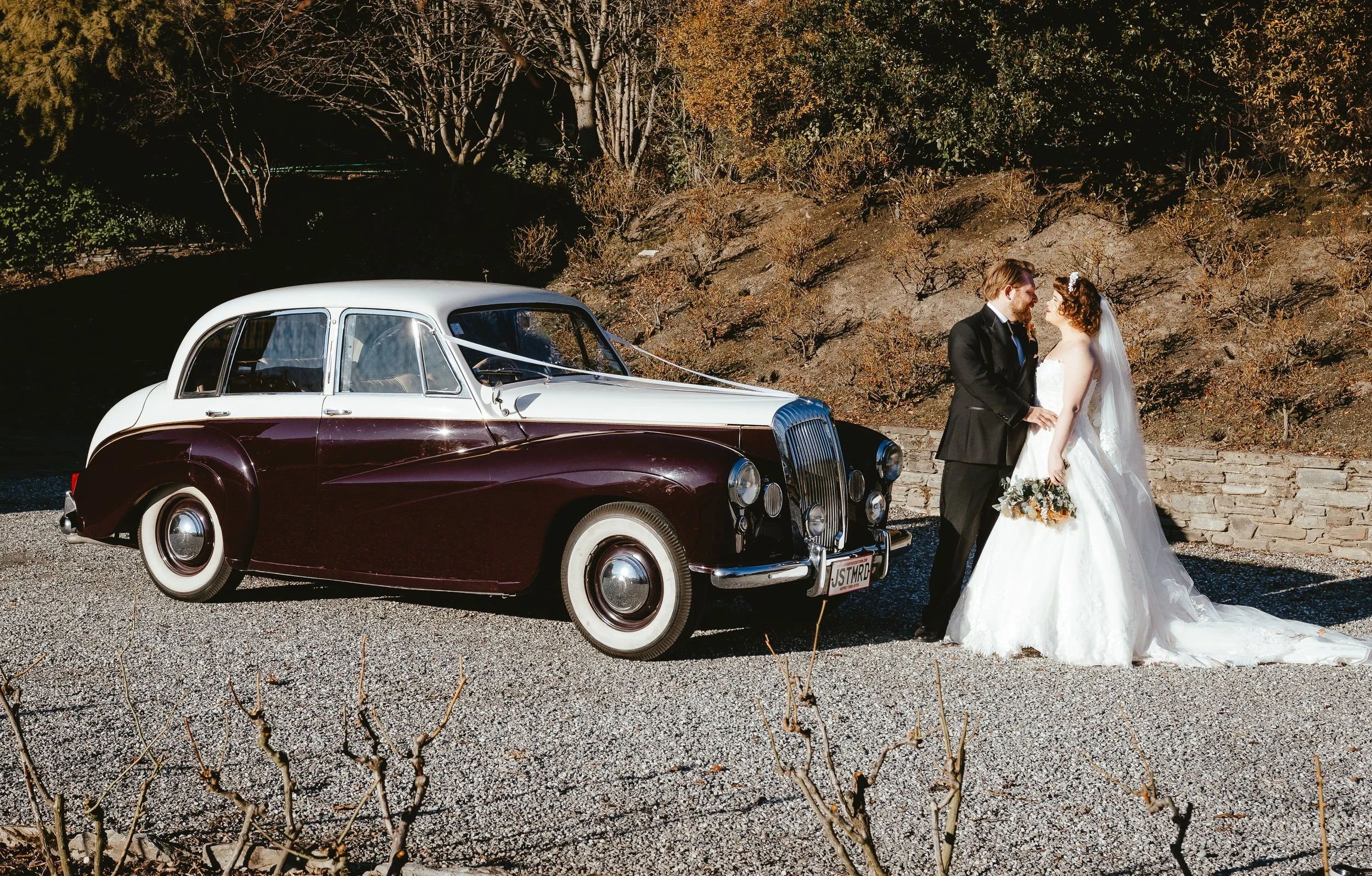 A bride and groom in wedding attire standing next to a vintage burgundy and white car, outdoors with trees and bushes in the background.