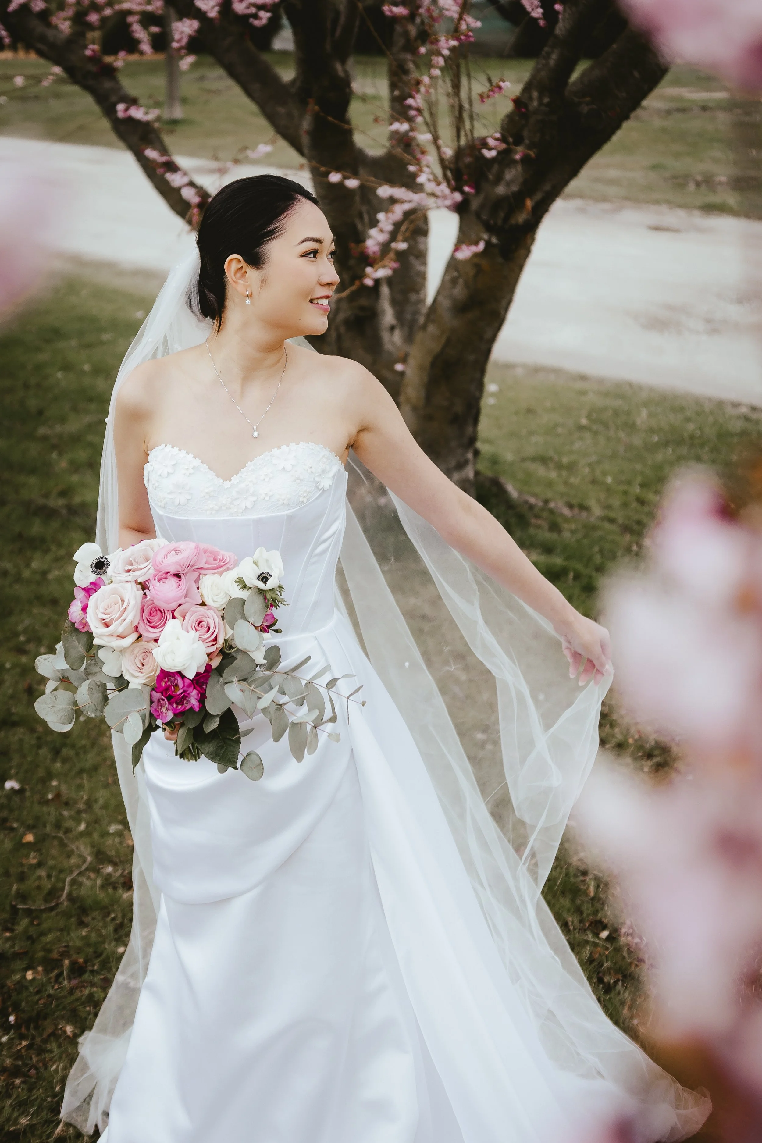 A bride in a white wedding dress holding a bouquet of pink, white, and purple roses, standing outdoors near a cherry blossom tree.