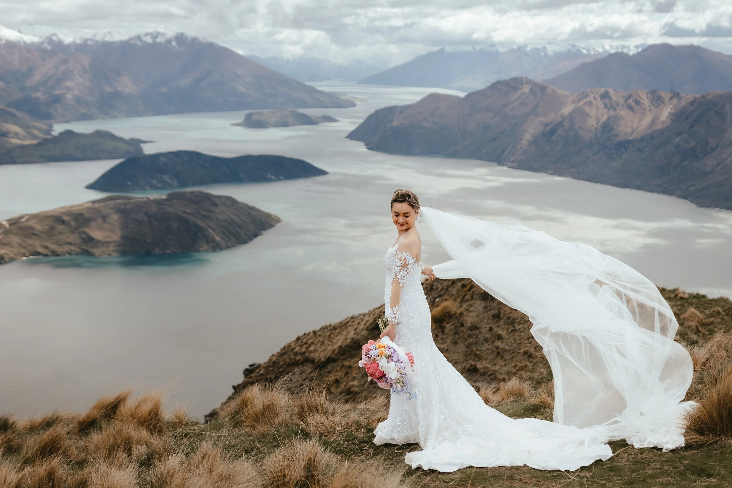 A bride in a wedding dress holding a bouquet stands on a hill with a lake and mountains in the background.