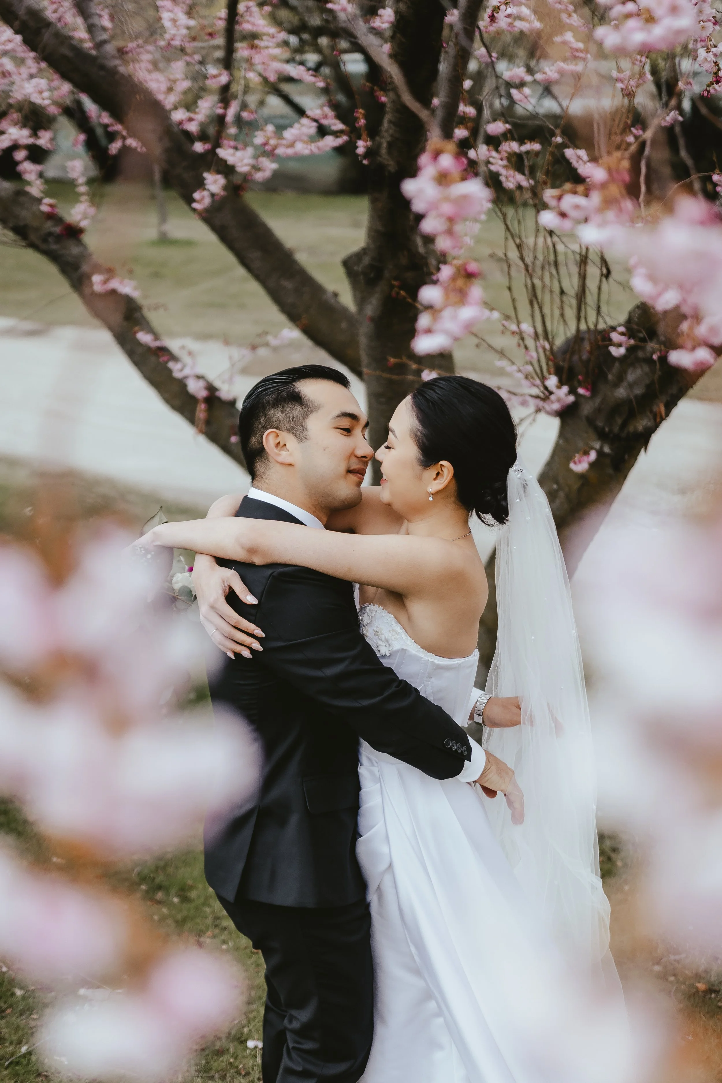 A bride and groom embrace and smile at each other in front of a blooming cherry blossom tree.