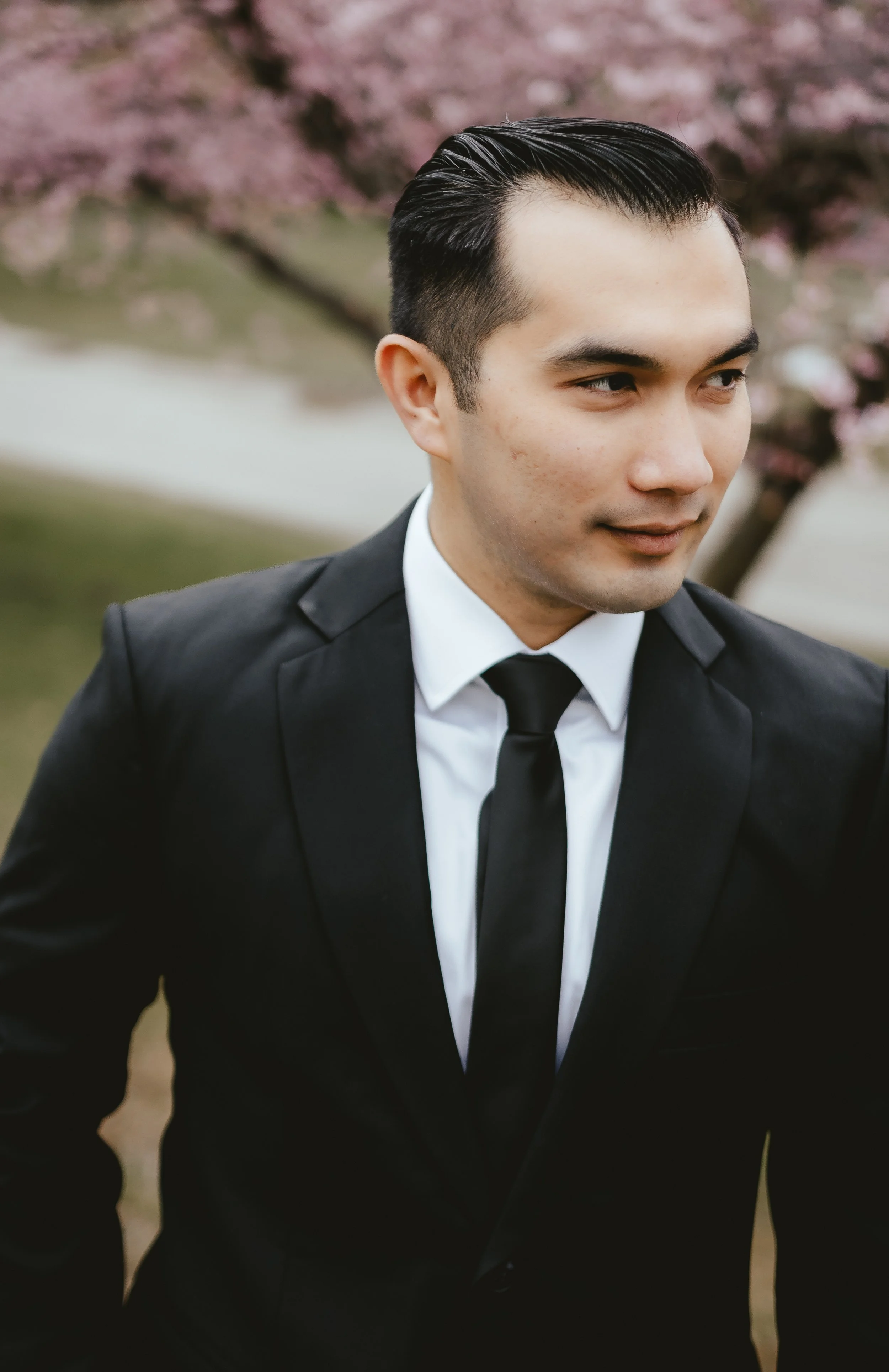 A man in a black suit with a white shirt and black tie outdoors, with a background of pink cherry blossom trees.