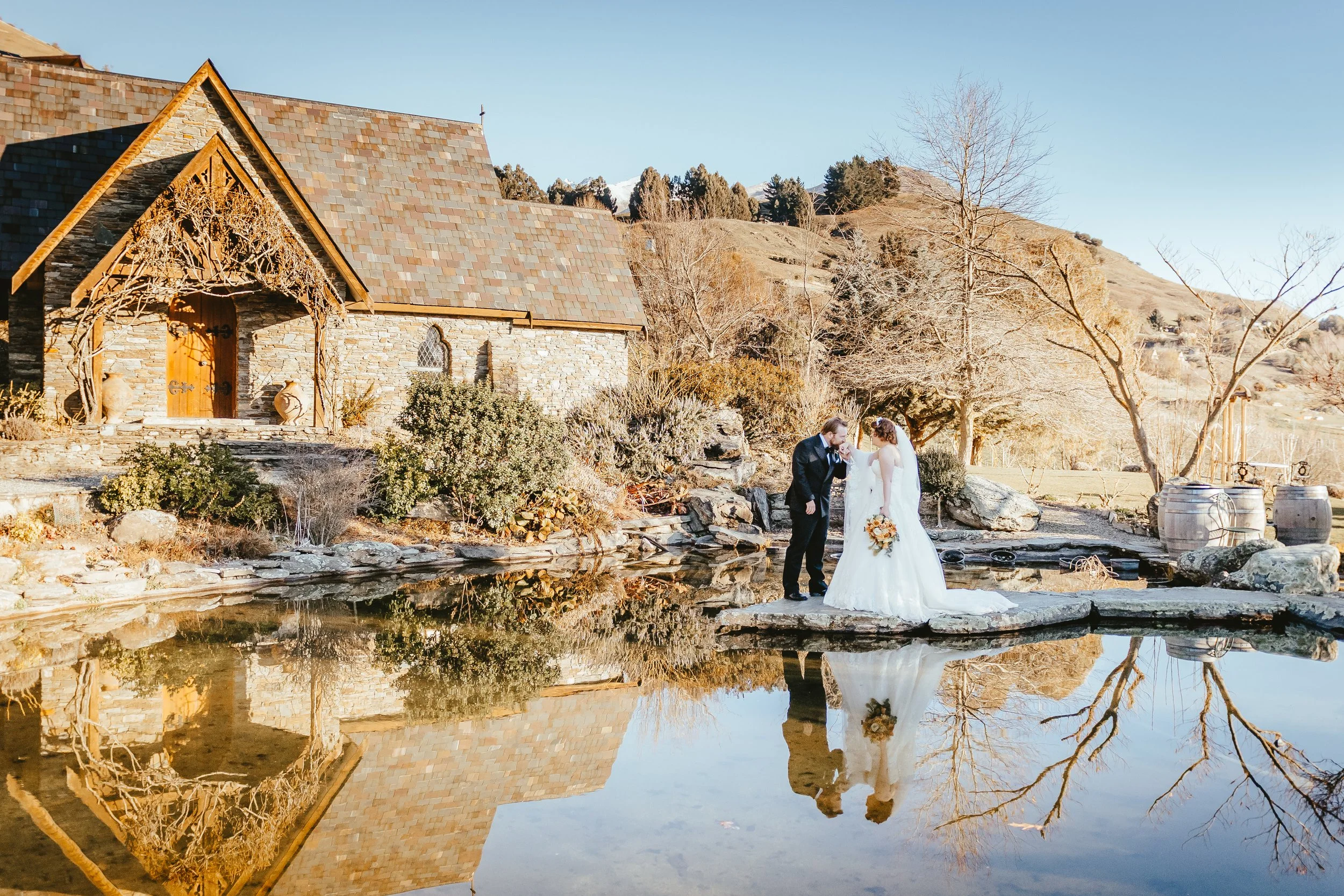 A bride and groom holding hands by a pond in a rural setting with a stone building, leafless trees, and rolling hills in the background. The scene is during late fall or winter with clear skies.