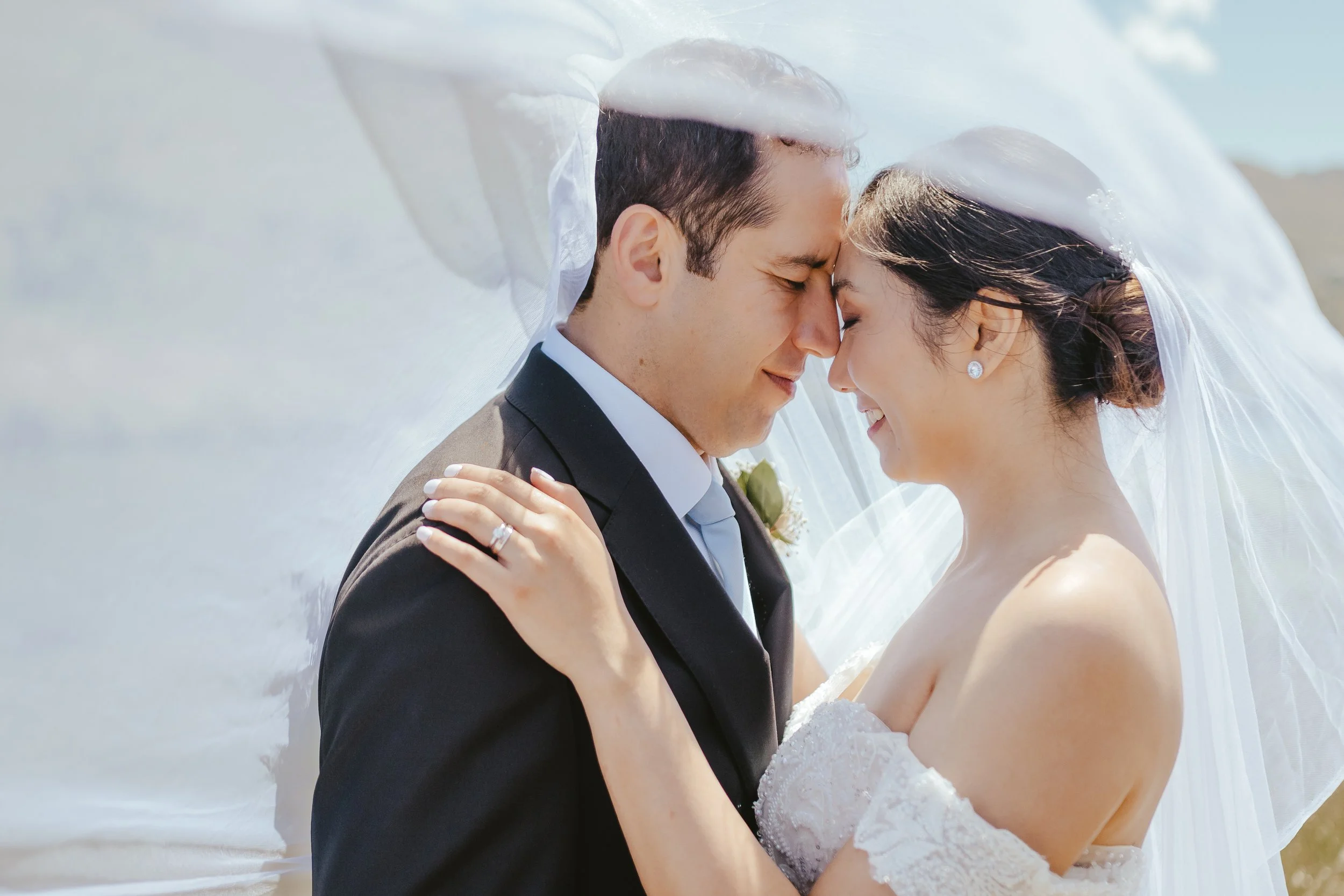 A bride and groom are lying outdoors with their foreheads and noses touching, their eyes closed, sharing an intimate moment under a flowing white veil on a sunny day.