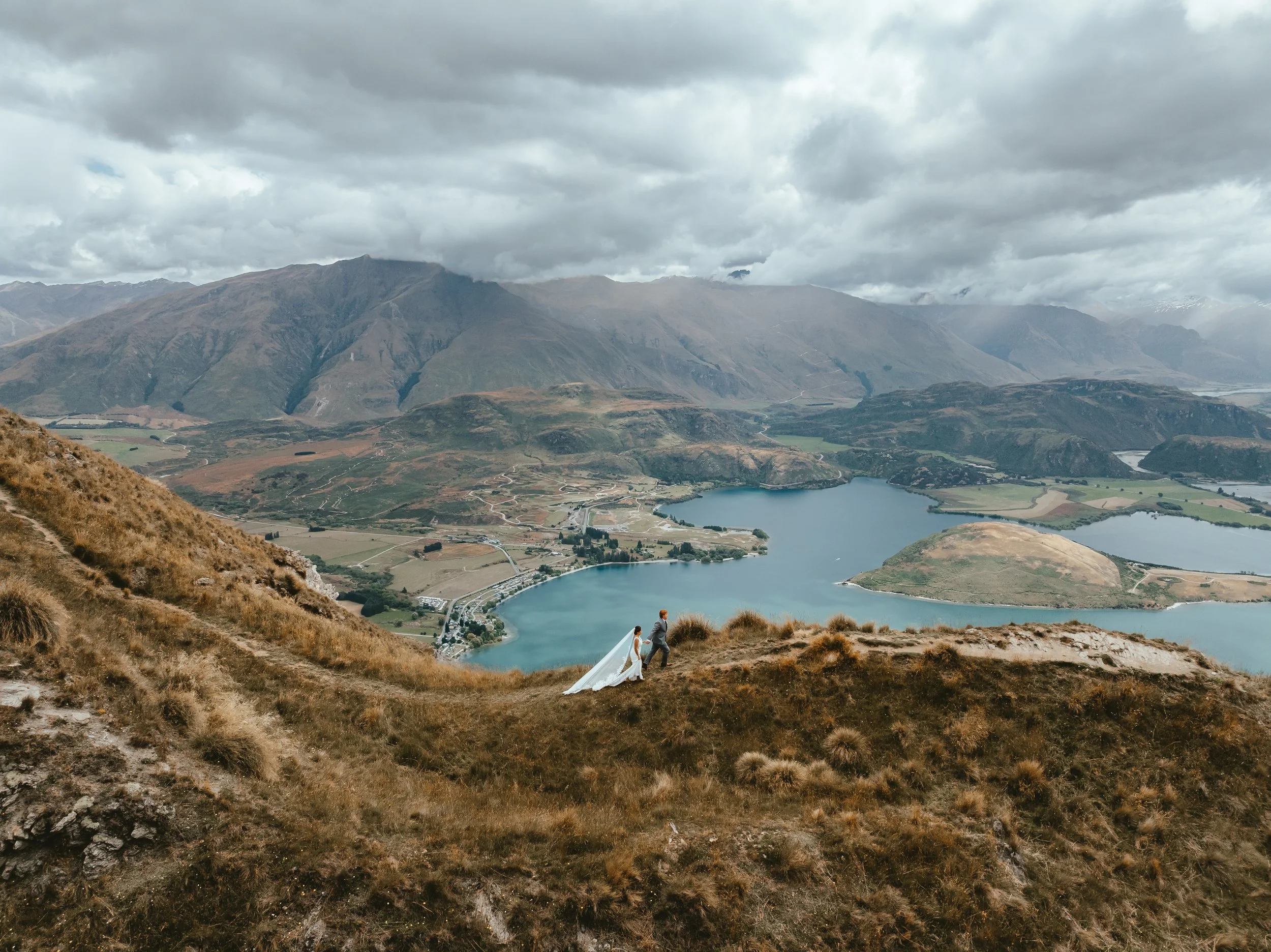 A bride and groom holding hands on a hillside with a lake and mountains in the background, under cloudy skies.
