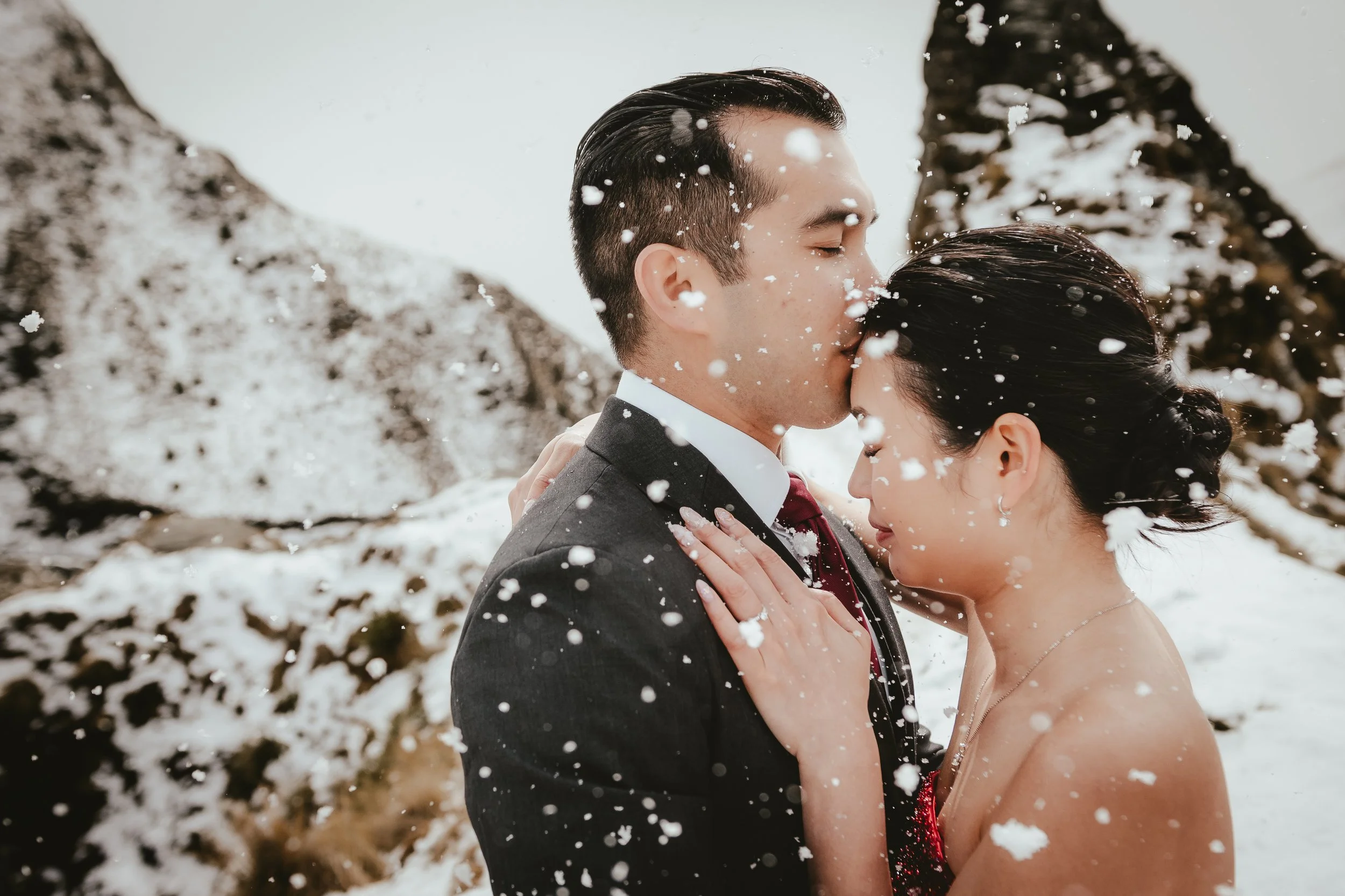 A couple dressed in formal attire embracing in a snowy outdoor setting, with snow falling around them.