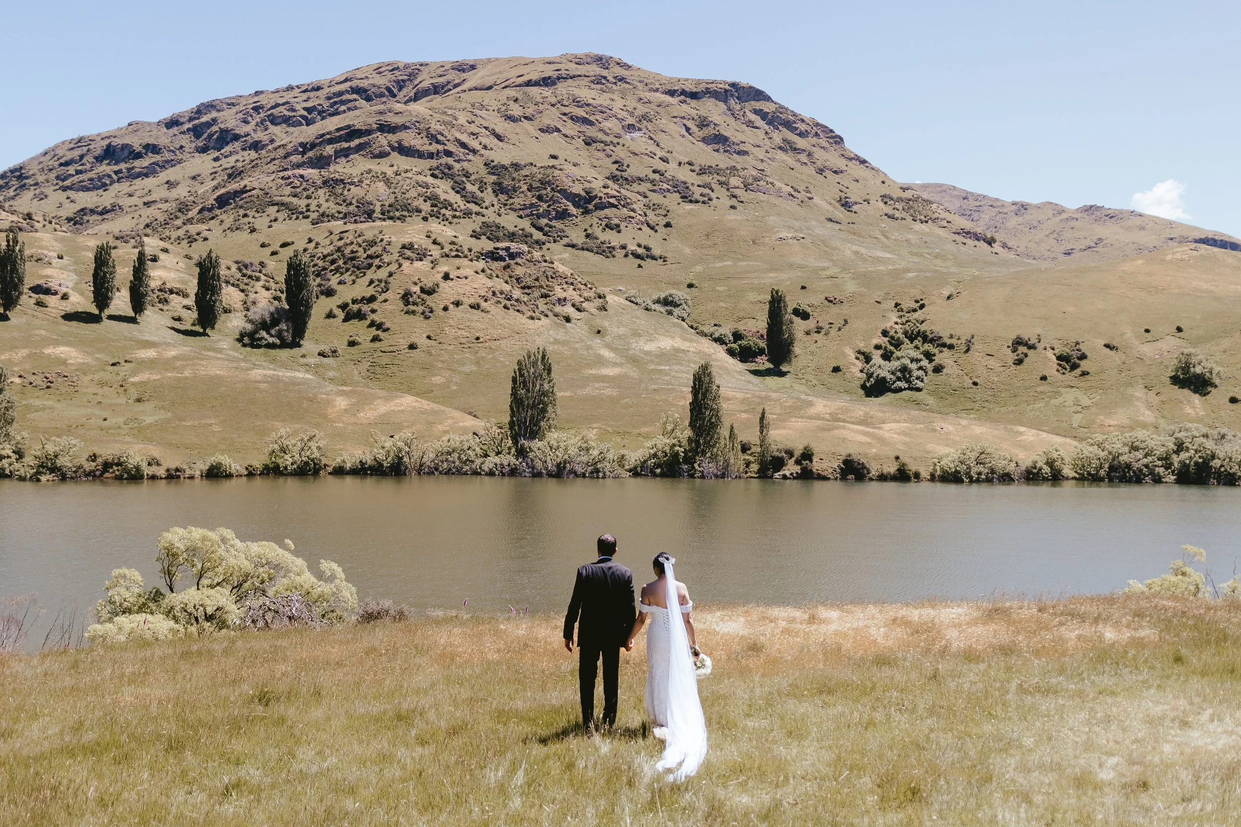 A bride and groom holding hands and walking towards a lake with a mountain in the background.