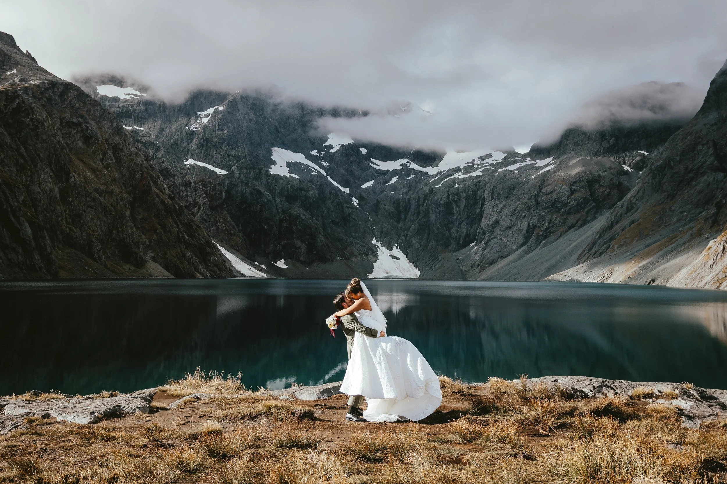 A bride and groom sharing a kiss beside a lake with mountain peaks and snow patches in the background.