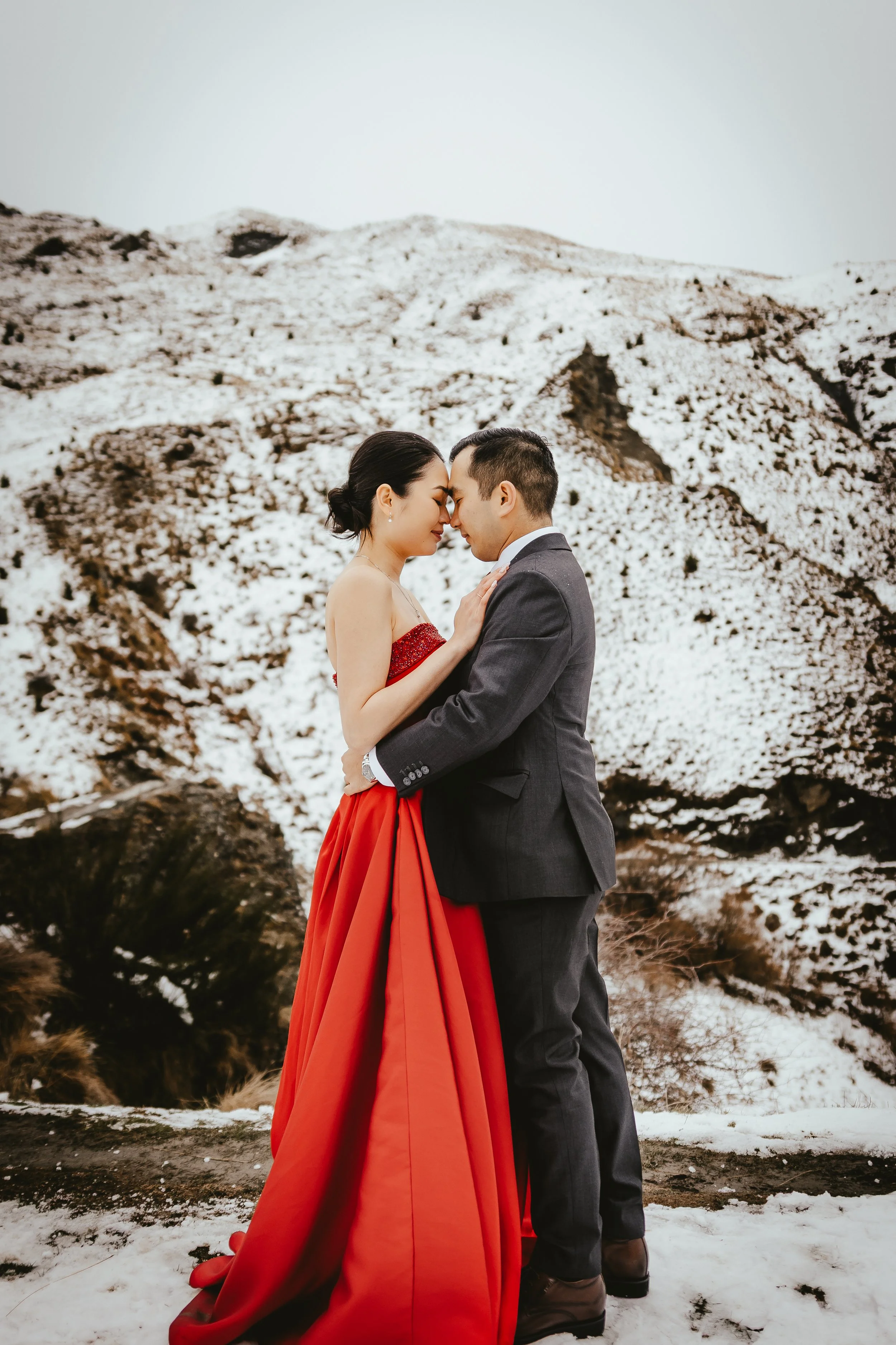 A couple embracing in a snowy outdoor landscape, with snow-covered mountains and trees in the background, the woman is wearing a red gown and the man a dark suit.