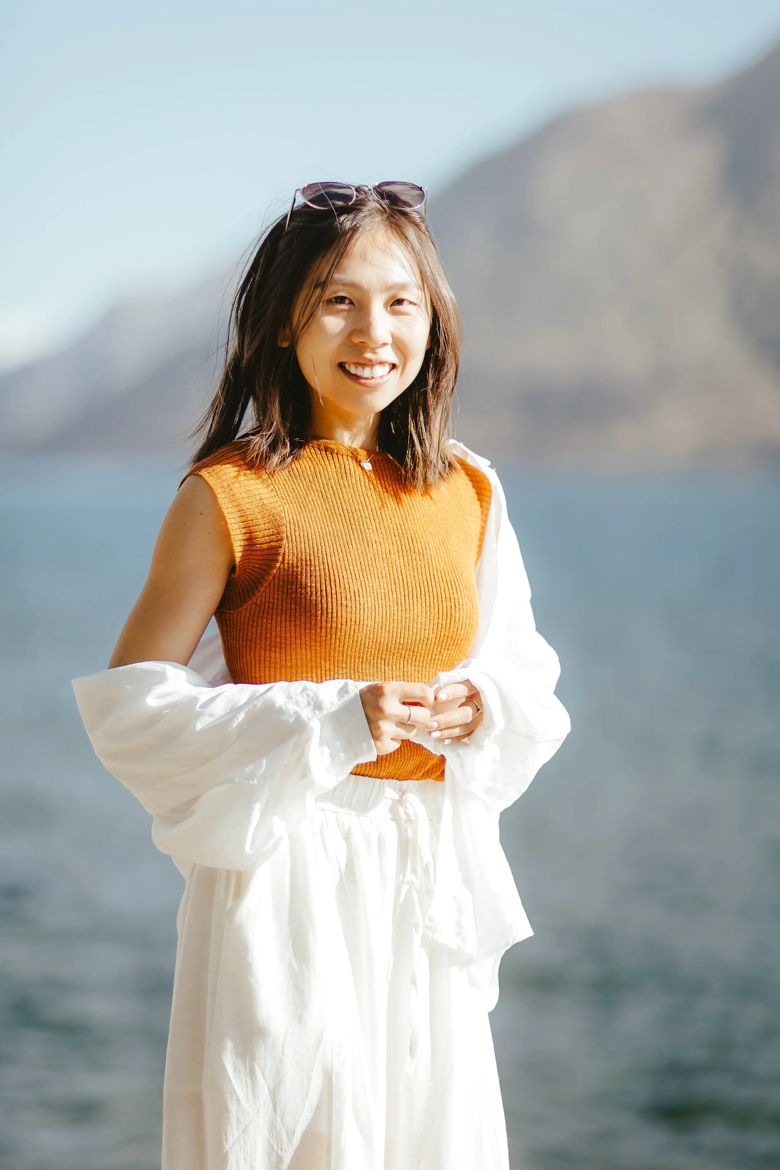 A woman with shoulder-length brown hair, sunglasses on her head, smiling and wearing an orange sleeveless top with a white jacket and white pants, outdoors near water with mountains in the background.