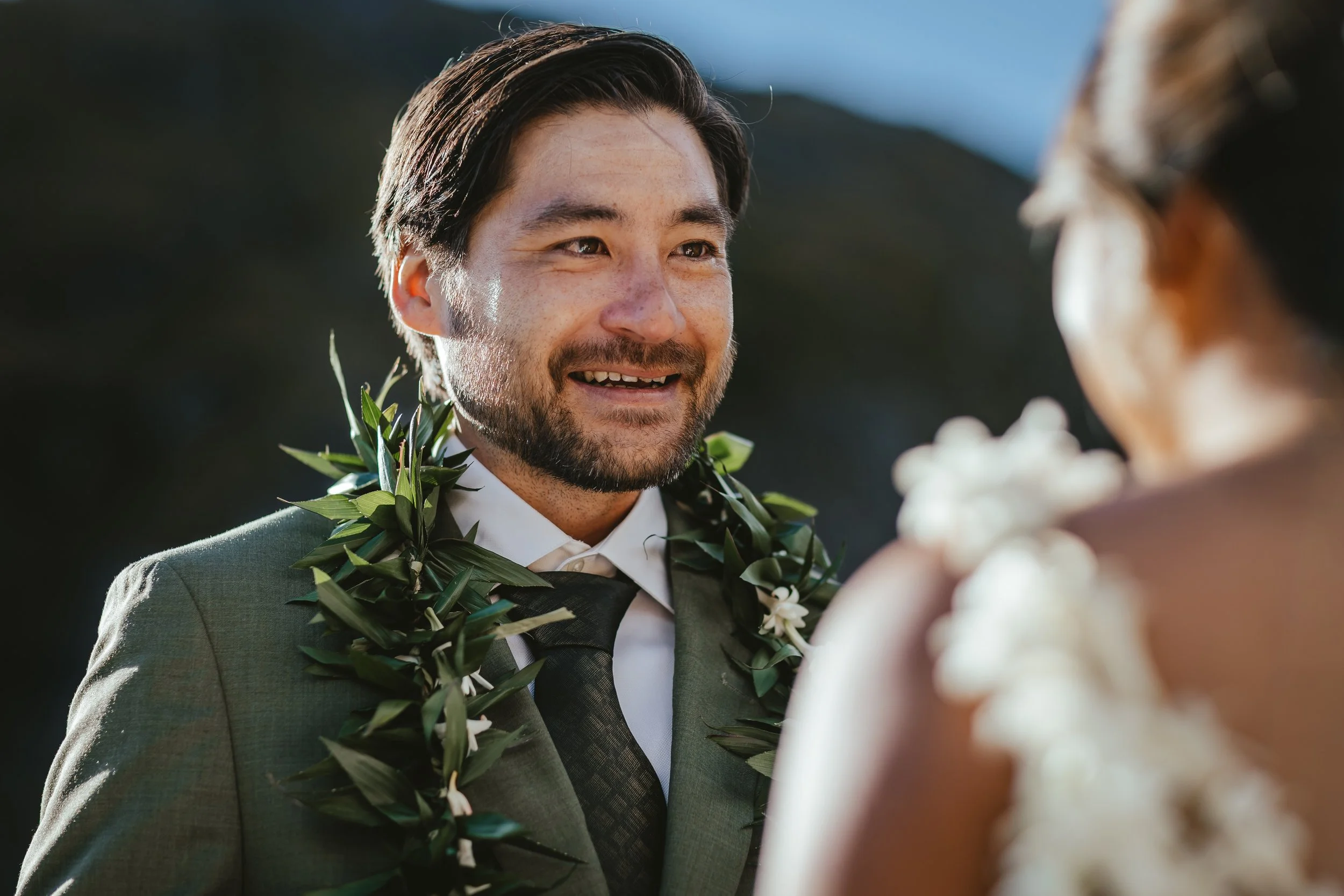 A smiling man wearing a gray suit and a green leafy lei, in a close-up outdoor setting during daytime, engaged in a conversation with someone whose shoulder and hair are visible in the foreground.