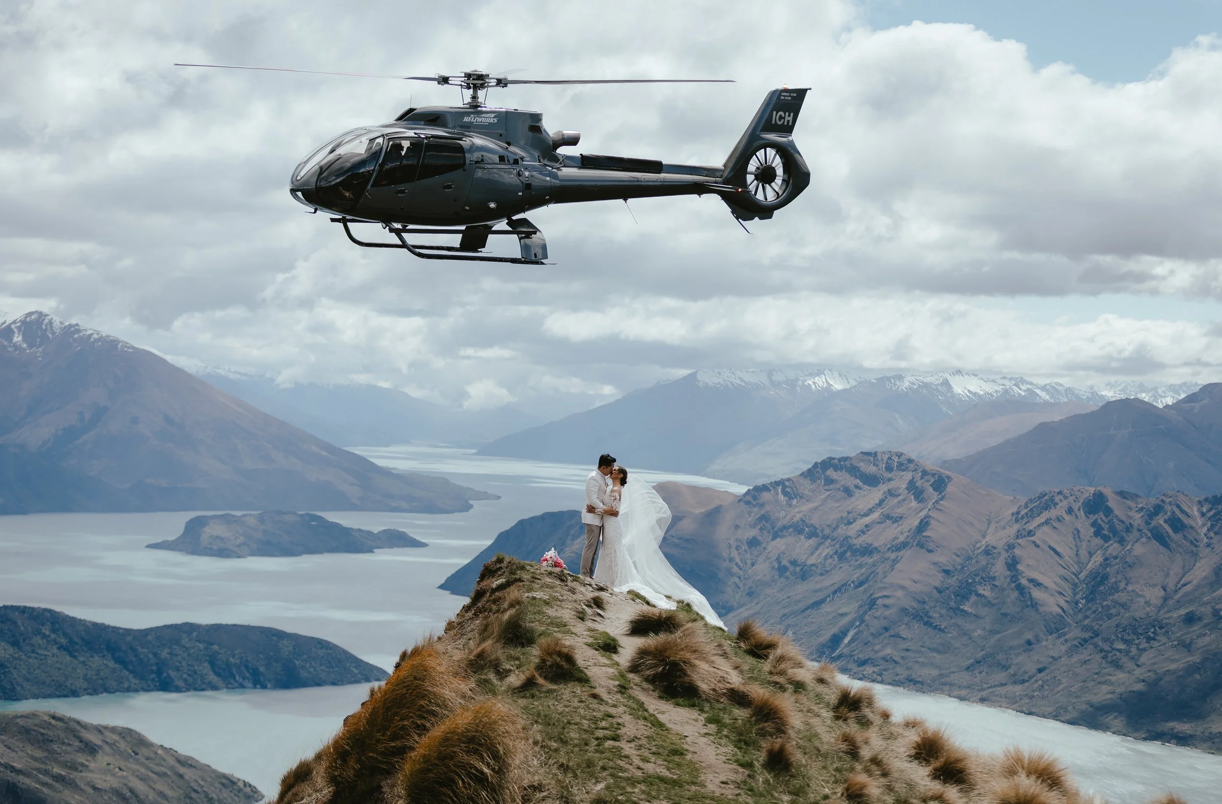A helicopter flying over a mountain landscape with a couple standing on a small hill, dressed in wedding attire.