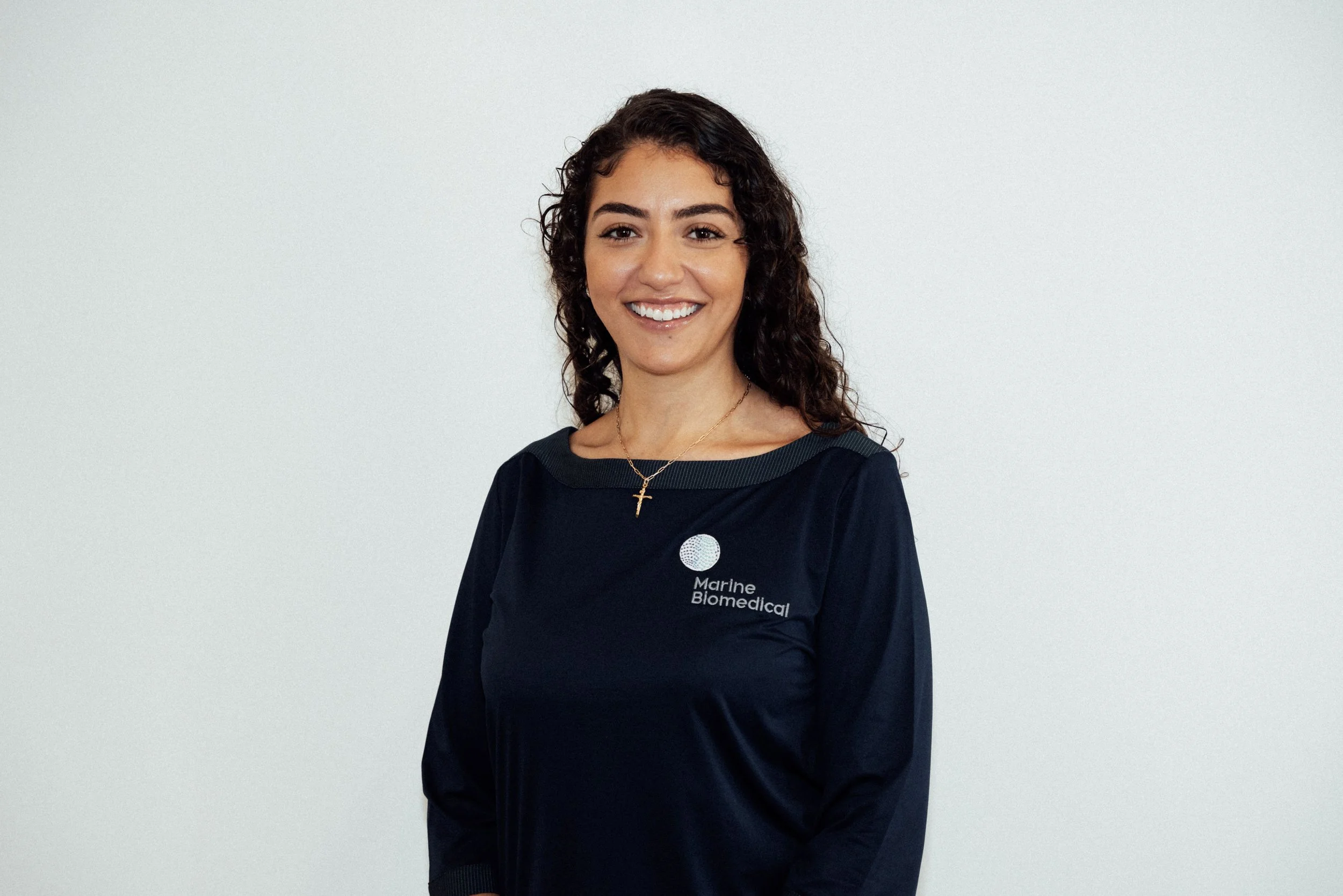 A young woman with curly brown hair smiling, wearing a black uniform with 'Marine Biomedical' written on it, standing against a plain light-colored background.