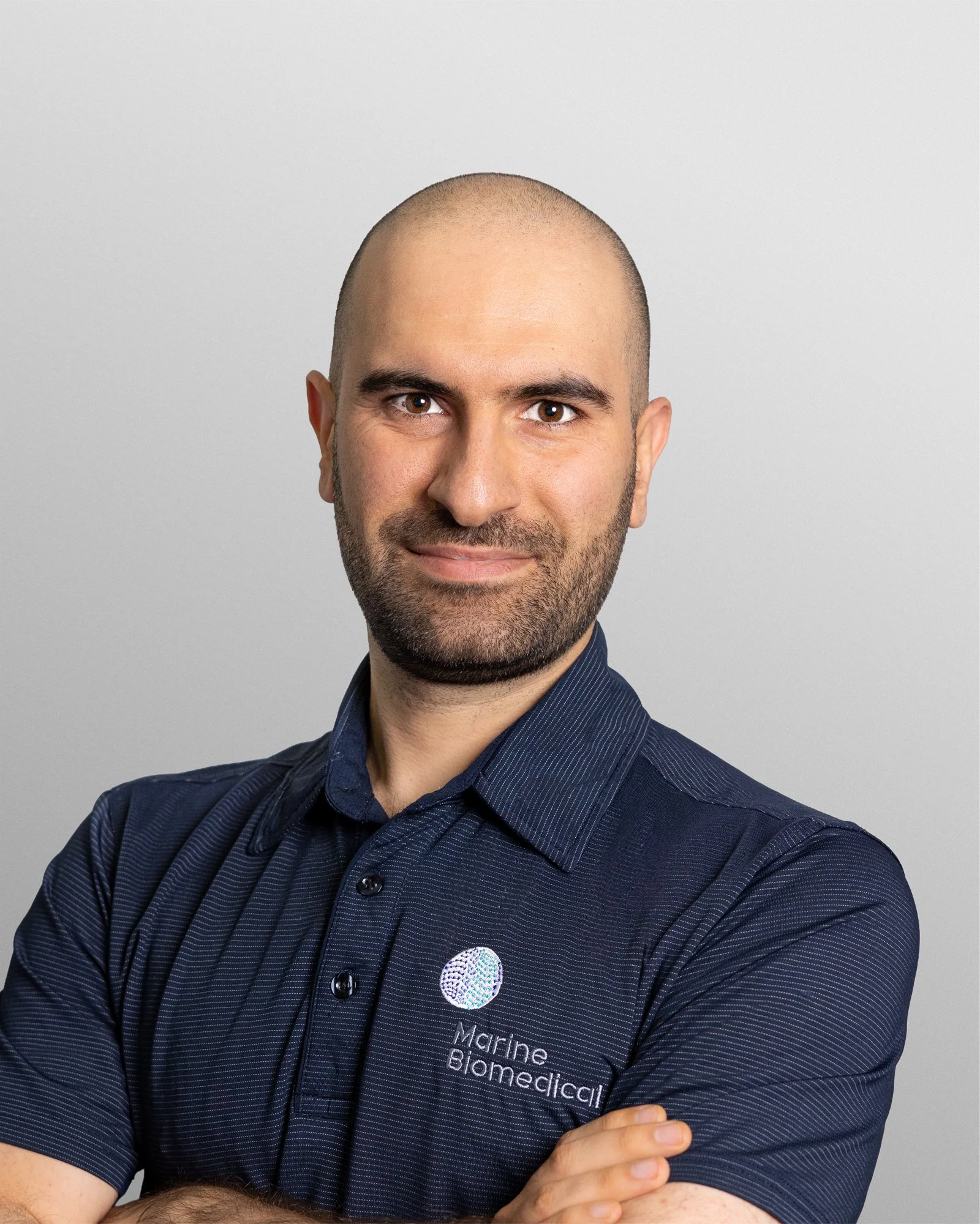 Portrait of a man with a shaved head and beard, wearing a dark blue shirt with the Marine Biomedical logo, smiling and standing against a plain gray background.