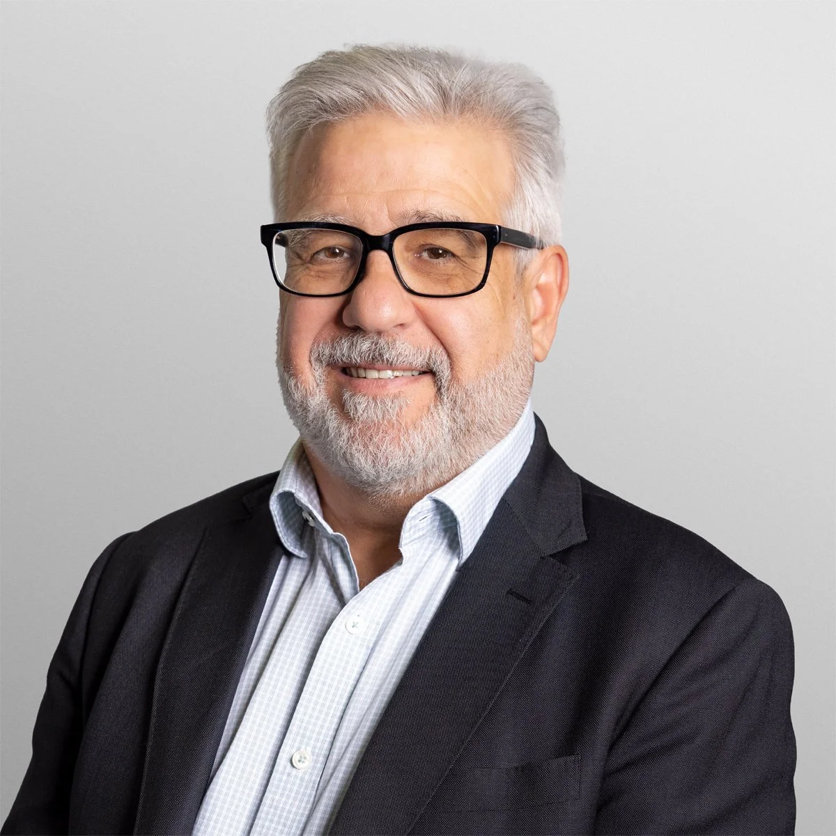 Headshot of a smiling older man with gray hair, beard, and glasses, wearing a black suit jacket and a light blue collared shirt against a plain light background.