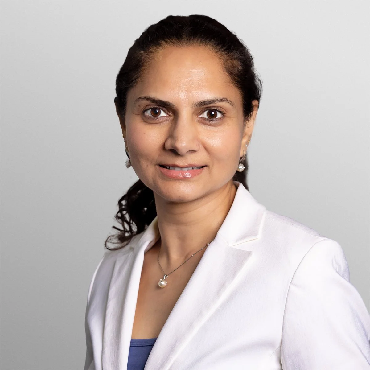 Headshot of a woman with dark brown hair, light skin, wearing earrings, a pearl necklace, and a white blazer against a plain light gray background.