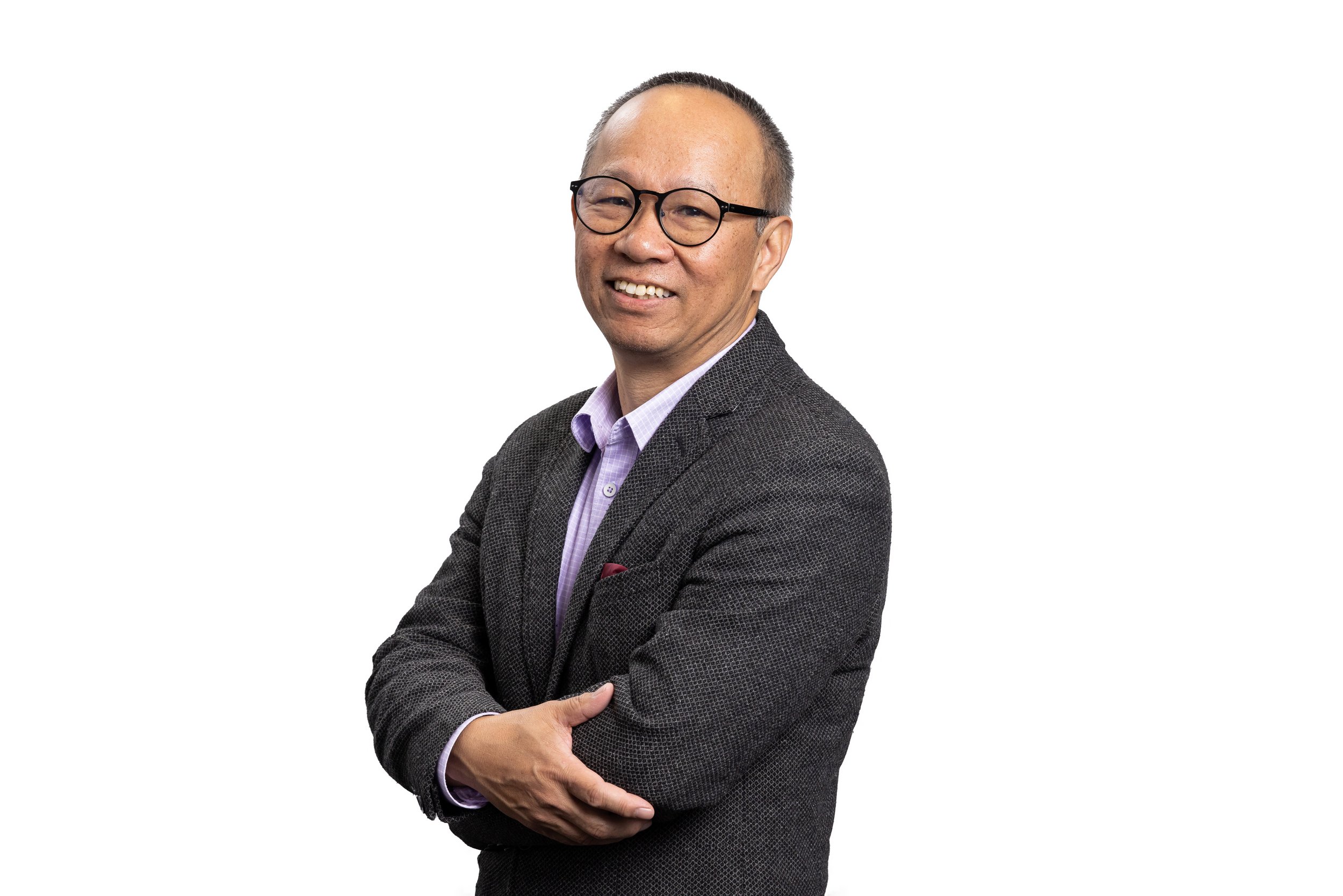 Portrait of a middle-aged Asian man wearing glasses, a gray blazer, and a purple shirt, smiling with arms crossed, against a white background.