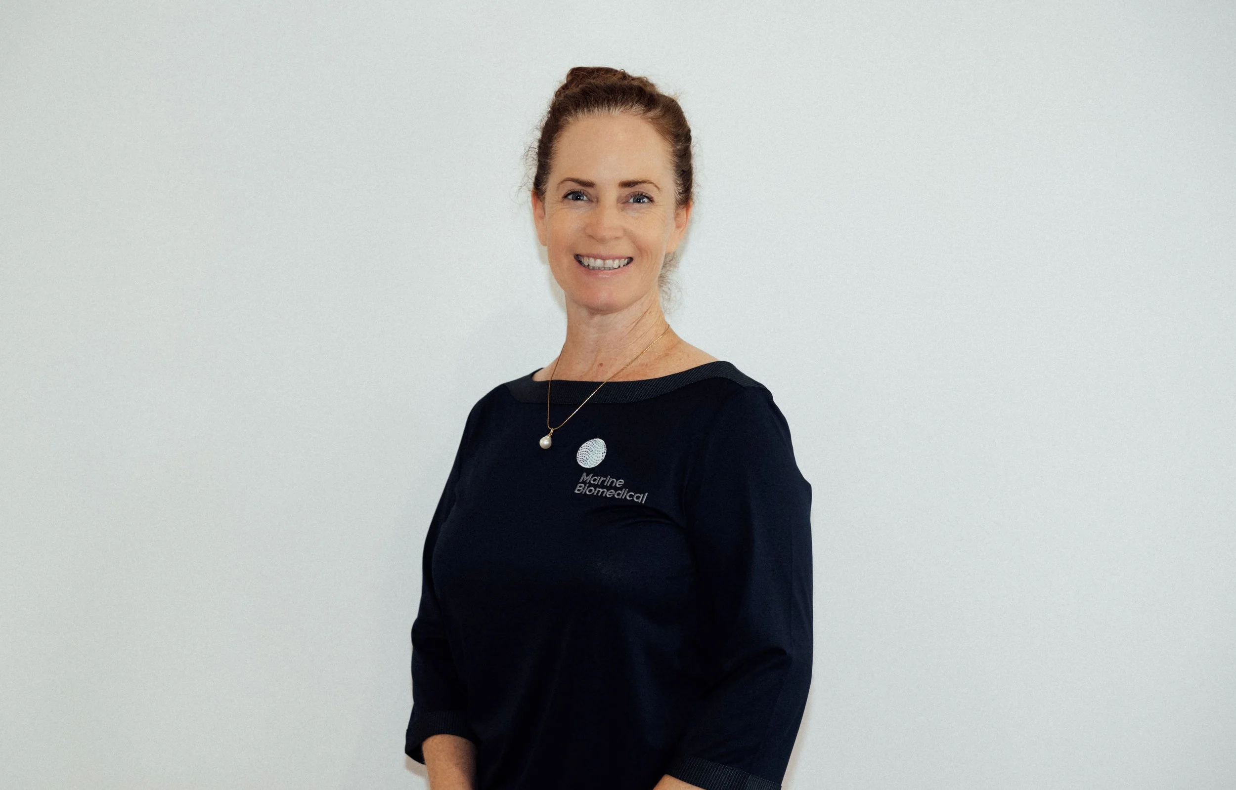 Smiling woman with brown hair styled in a bun, wearing a black Marine Biomedical shirt, standing against a plain white wall.