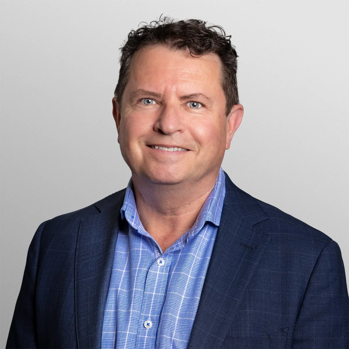 A professional headshot of a middle-aged man with short, curly dark hair, blue eyes, wearing a blue checkered shirt and a dark blue blazer, standing against a plain light gray background.