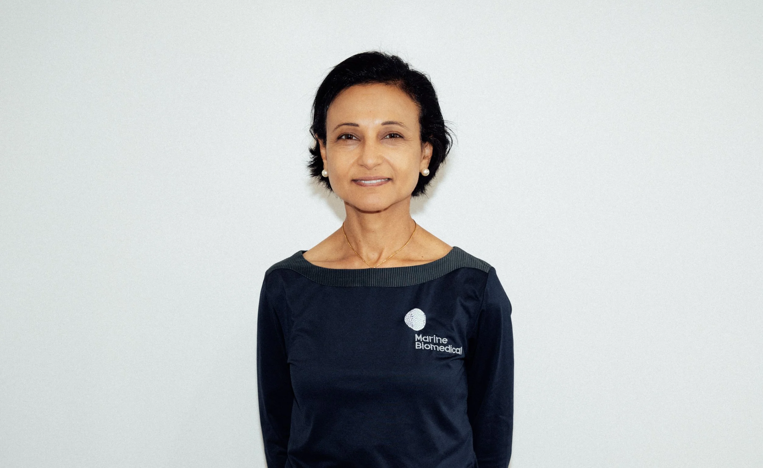 A woman with short black hair and earrings wearing a navy blue Marine Biomedical shirt standing against a plain white wall.