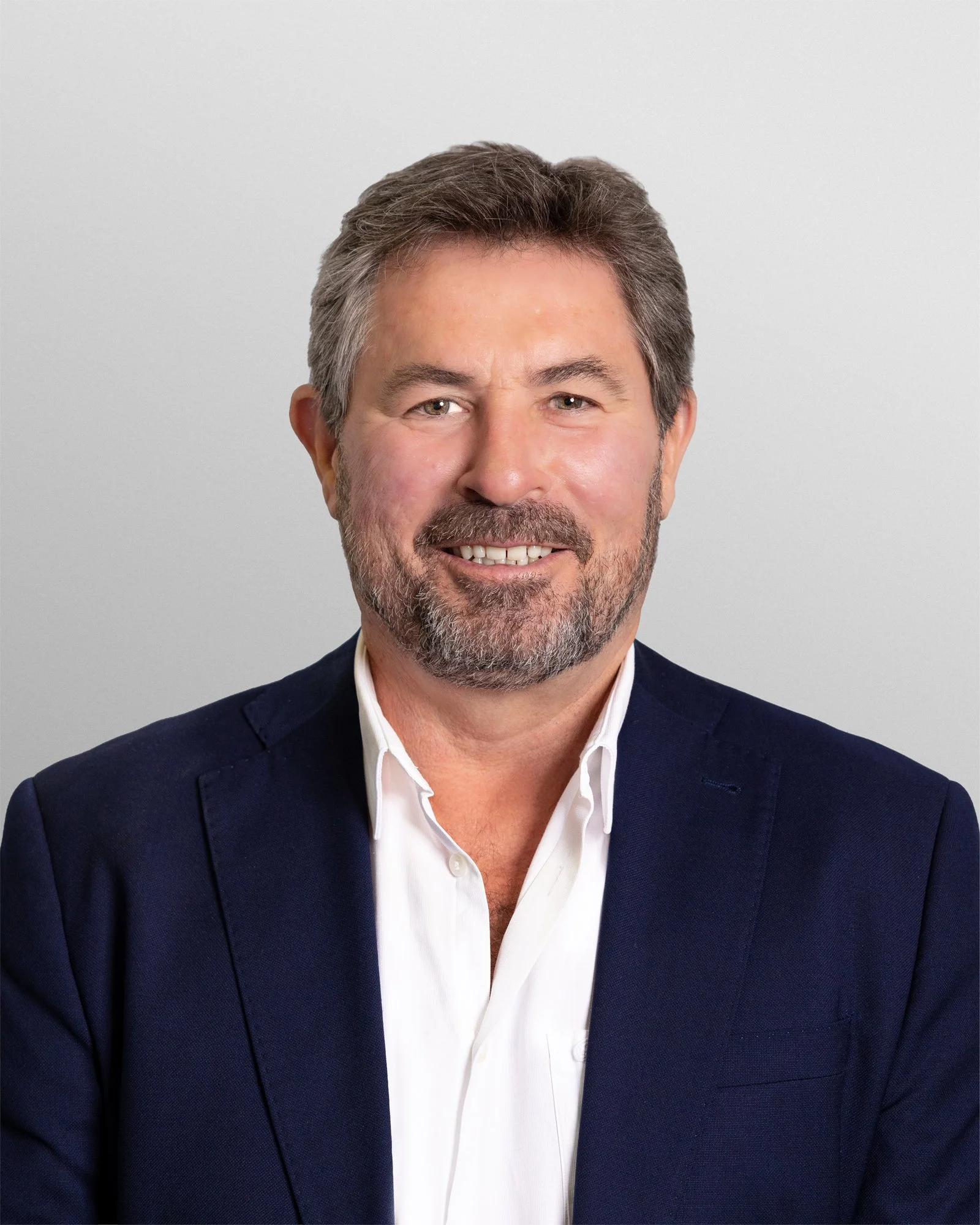A man with gray hair and beard smiling, wearing a suit jacket and white shirt, against a plain background.