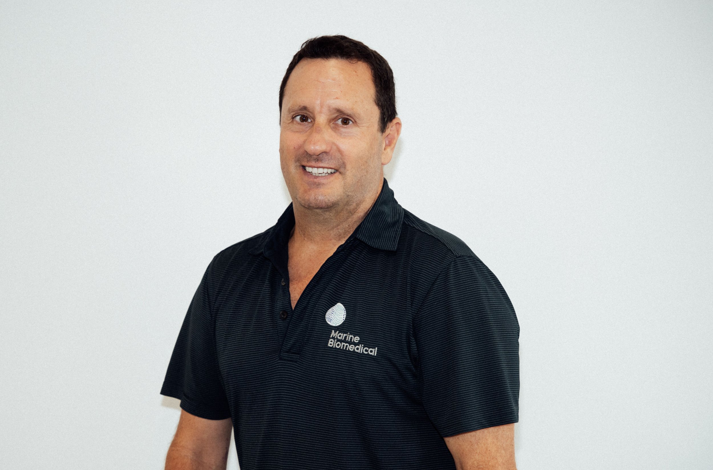 A man in a black polo shirt with a Marine Biomedical logo on it, standing against a plain white background, smiling at the camera.