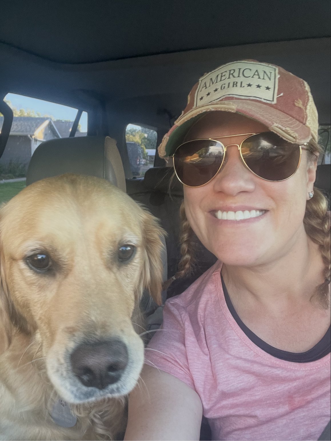 A woman wearing a pink shirt, sunglasses, and a distressed cap, smiling, sitting beside a golden retriever inside a vehicle, with houses visible through the windows.