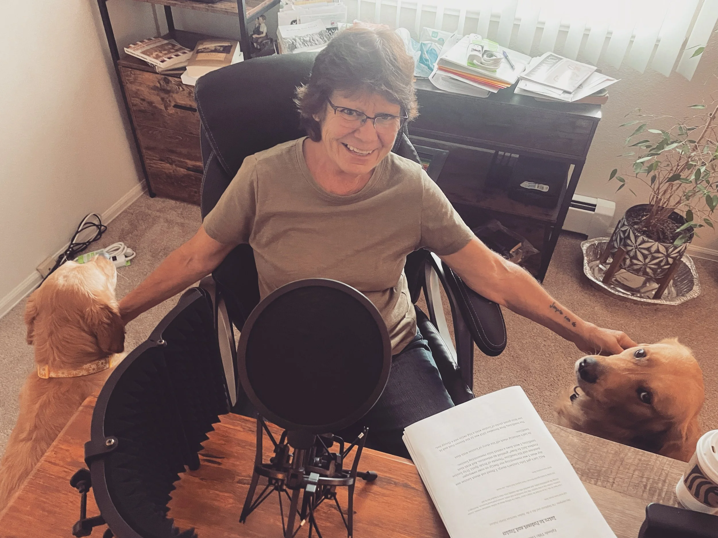 A woman with short brown hair and glasses smiling while sitting in a wheelchair, petting two golden retrievers. One dog is on her left side with a yellow bandana, and the other is on her right side, looking up at her. She is in a room with a wooden desk, a speaker, books, and a potted plant with a decorative tray on the floor.