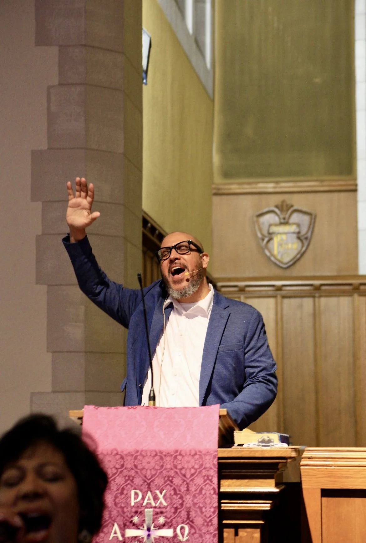 A man with glasses and a beard, wearing a blue blazer and white shirt, is speaking or singing at a podium inside a church or religious building. He is raising his right hand and appears to be passionately engaged. There is a woman in the foreground, partly visible, with her mouth open. Behind him, there is a pink cloth with religious symbols and the words "PAX A..."