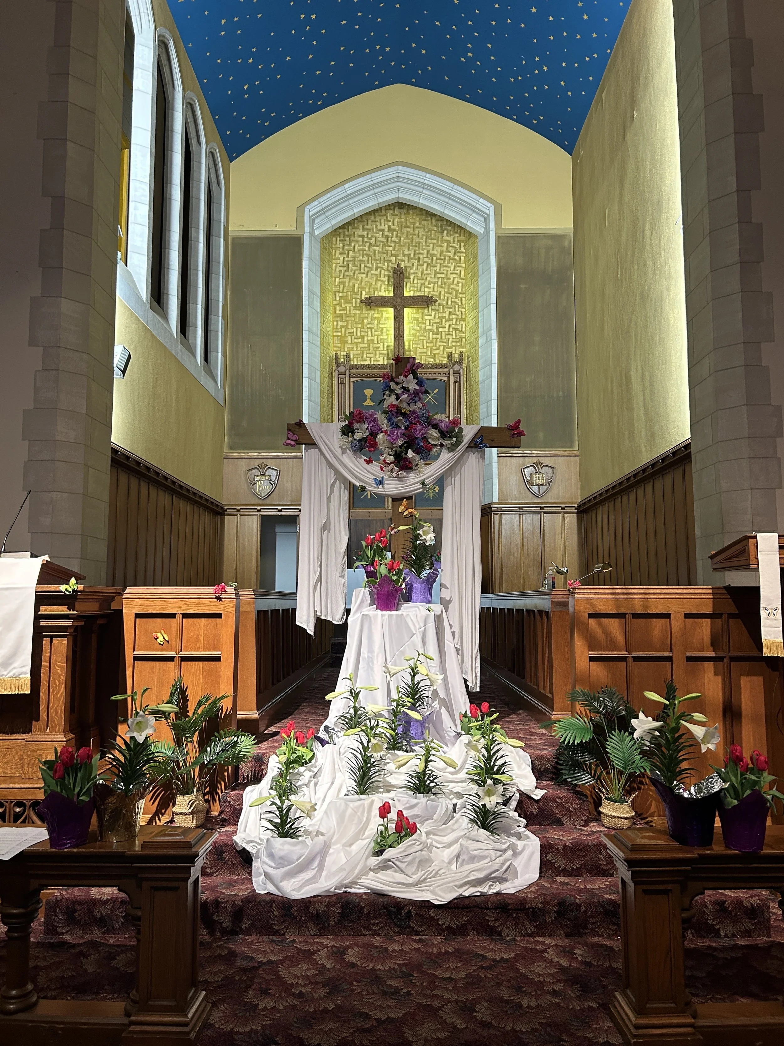 Inside a church sanctuary decorated with flowers for a wedding or religious ceremony, featuring a cross at the altar, floral arrangements, and draped fabric.