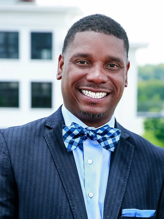 A smiling man wearing a dark pinstripe suit, light blue shirt, and a blue checkered bow tie, standing outdoors in front of a modern building.
