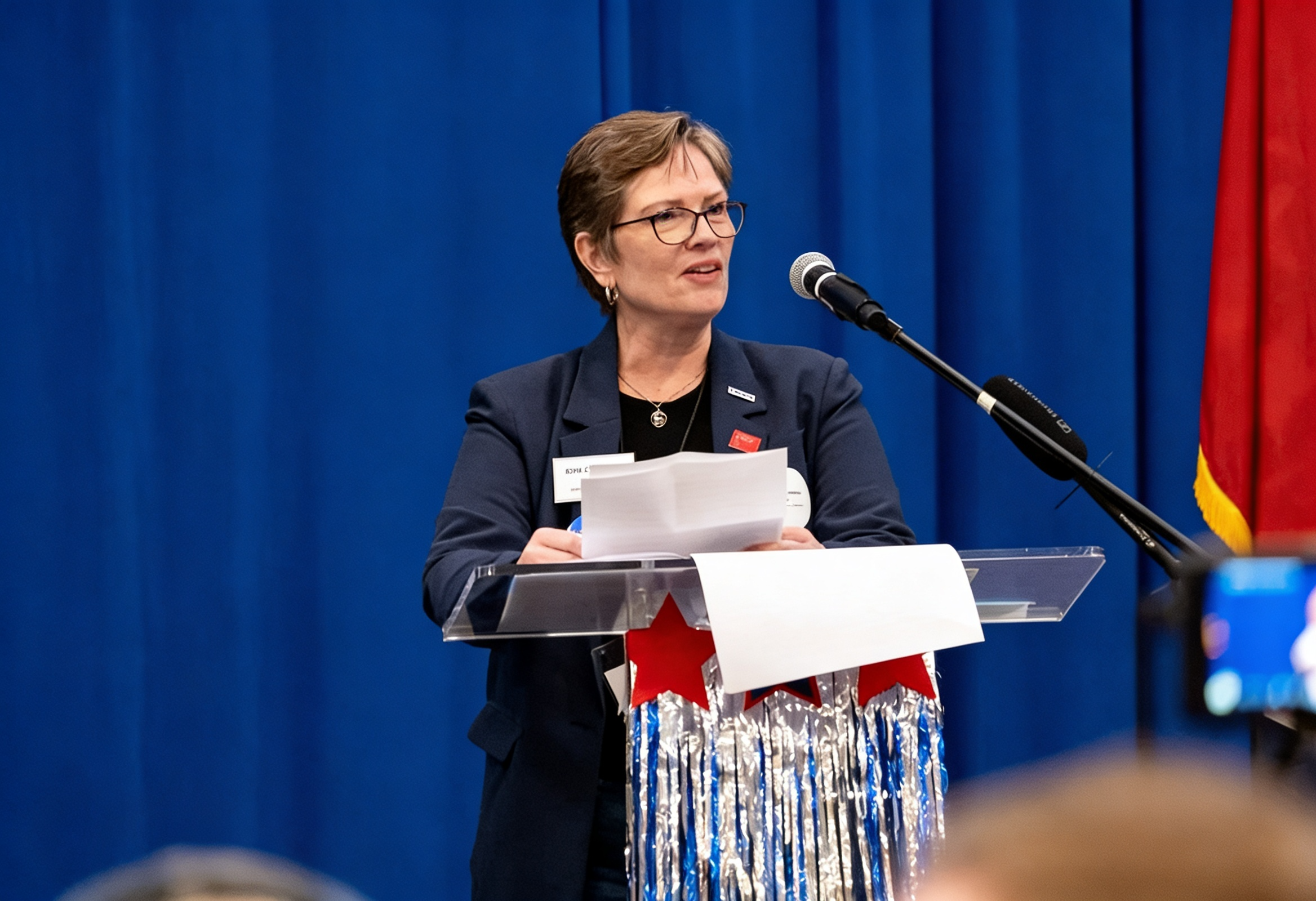 A woman with short hair, glasses, and earrings speaking at a podium with a microphone, in front of blue and red curtains, holding papers. There are American flags and a sign with her name on the podium.