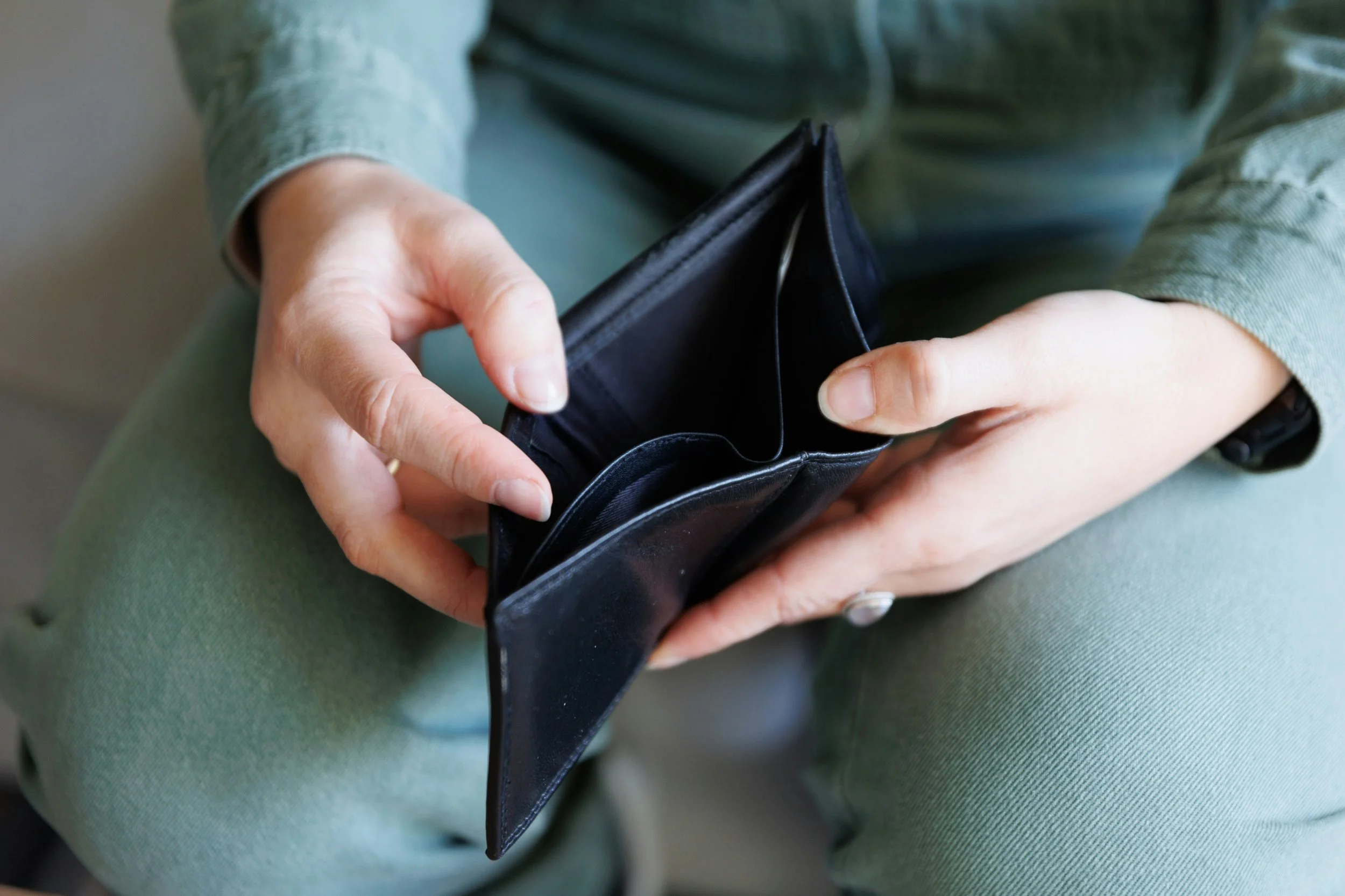 A person in a long-sleeve green shirt is holding an open empty black wallet over their lap, which is covered in green fabric.
