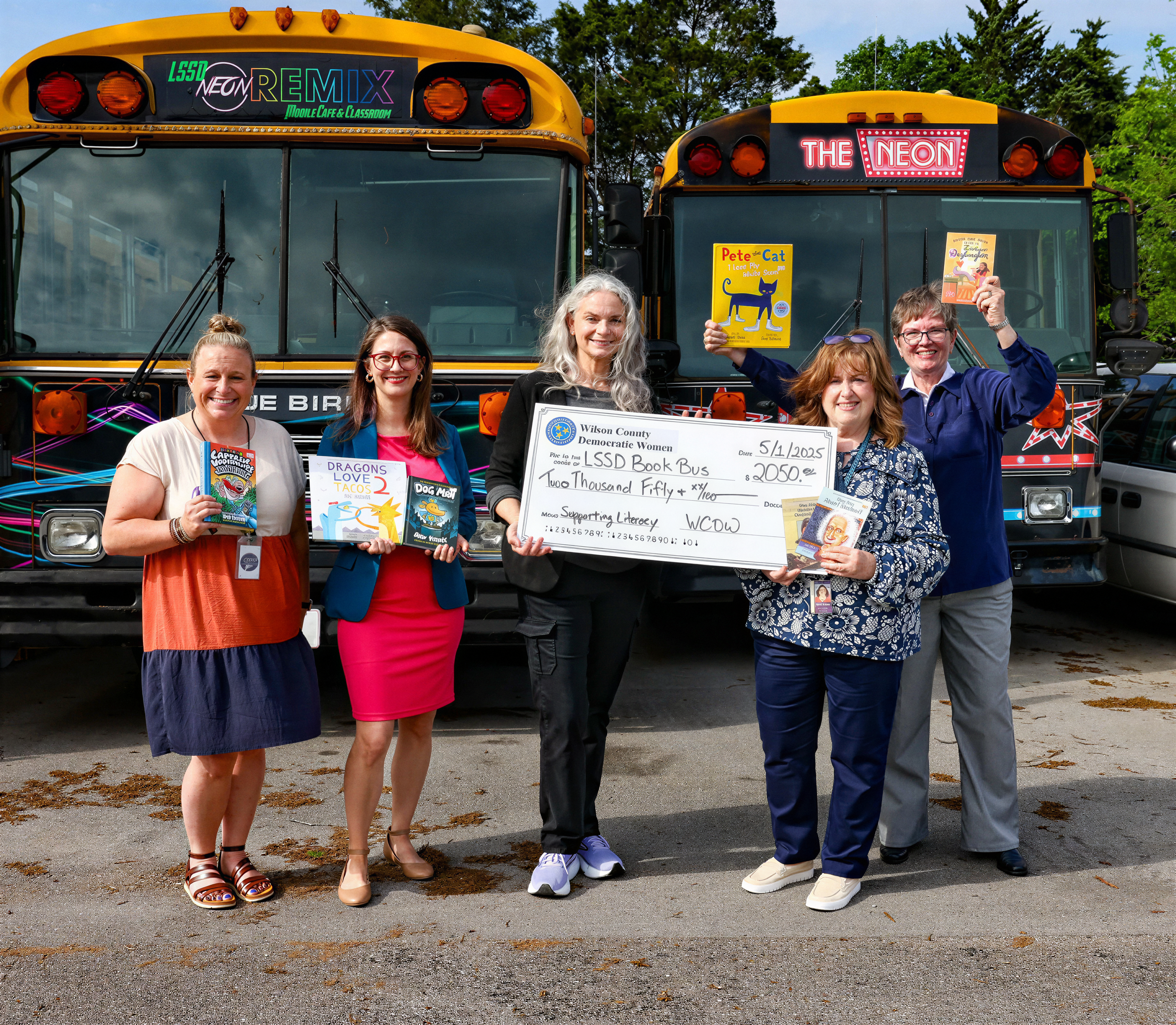 Five women standing in front of two school buses holding books, checks, and signs. One bustles a dollar bill, one has books about dragons, tacos, and dog meat, and another holds a large ceremonial check from Wilson County Democratic Women. The women are smiling and dressed casually, with one wearing a floral shirt and others in colorful clothing. The buses have neon and sign graphics, and the scene appears to be outdoors on a partly cloudy day with trees in the background.