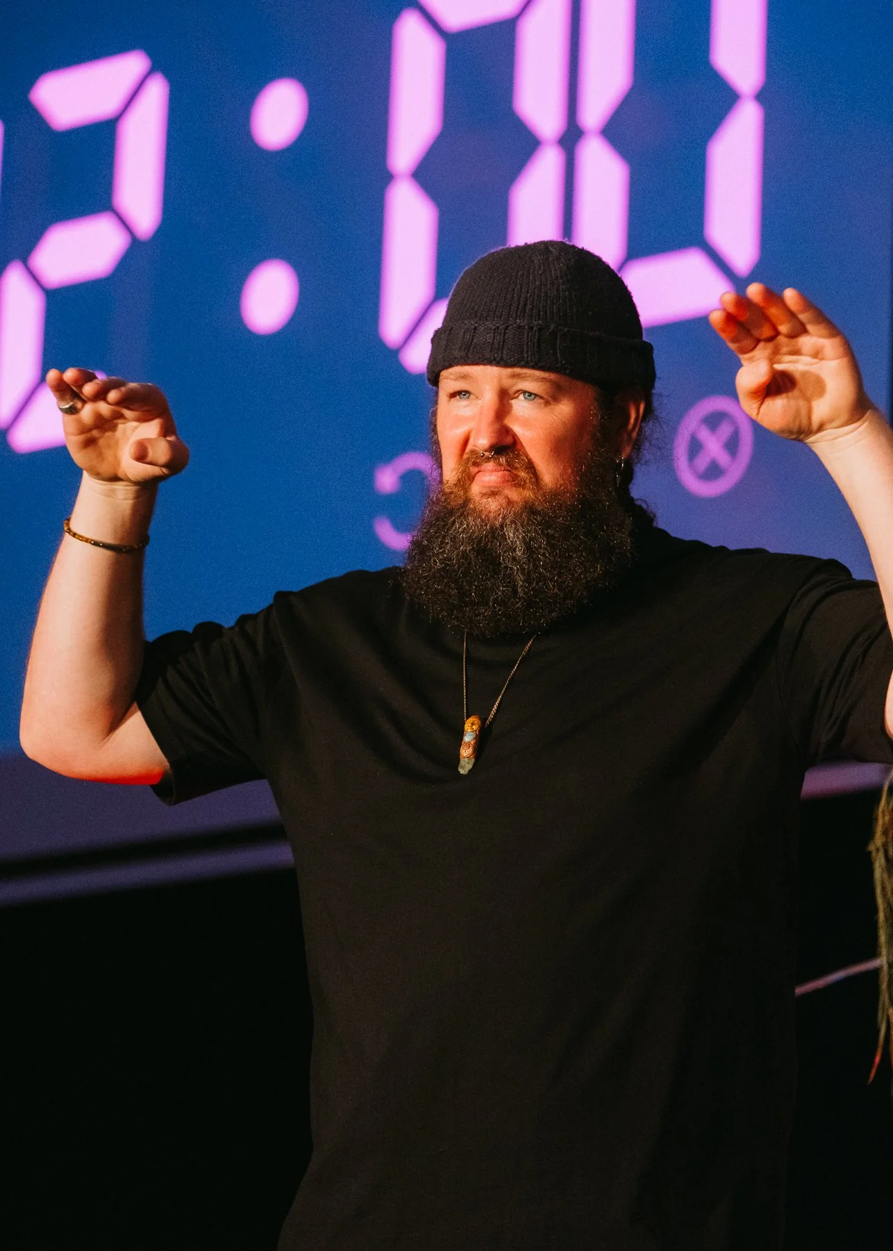 Man with a beard wearing a black beanie and black t-shirt, raising his hands near his head, with a digital purple clock display in the background.