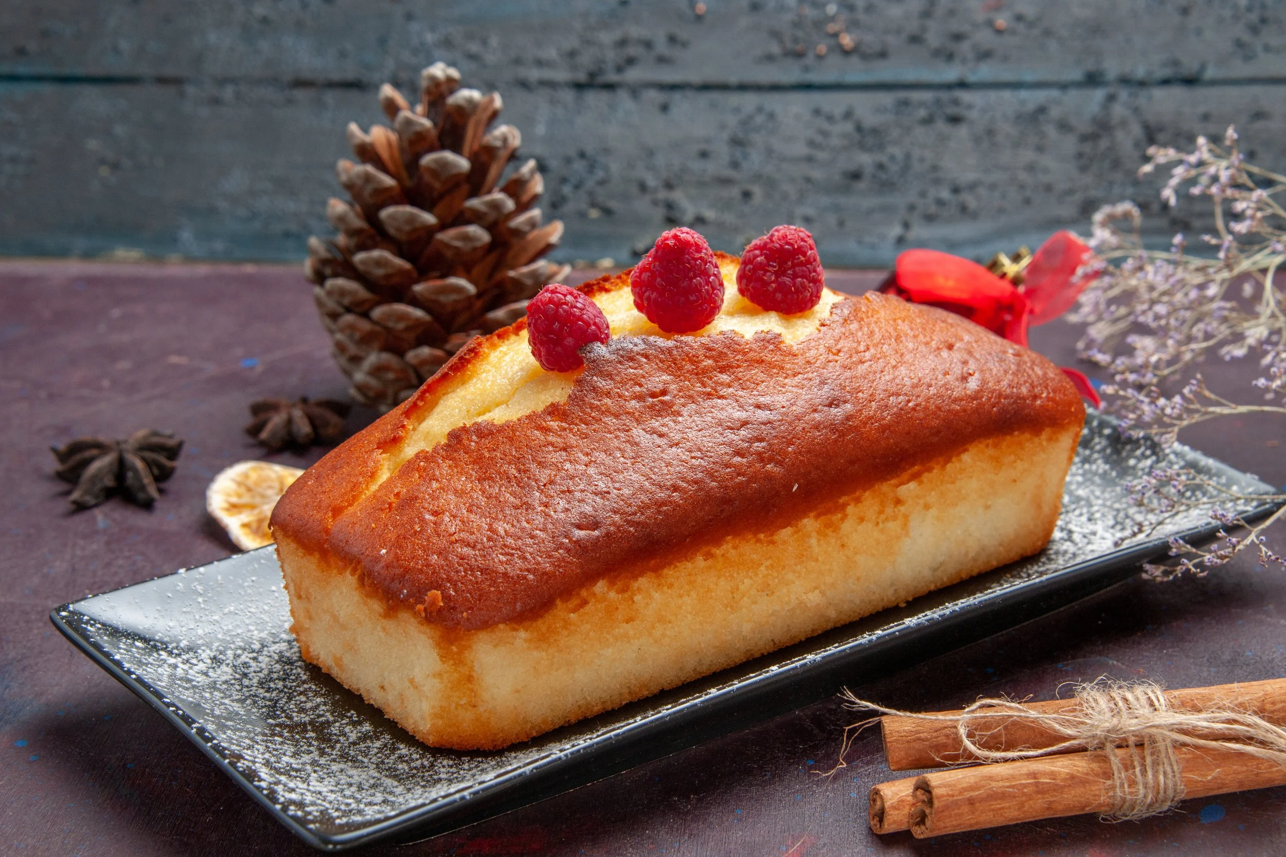 A rectangular sponge cake with a golden-brown top, topped with three raspberries, on a black rectangular plate dusted with powdered sugar. Surrounding the cake are a pine cone, star anise, dried citrus slices, a bundle of cinnamon sticks, a red flower, and sprigs of dried flowers, with a rustic dark background.