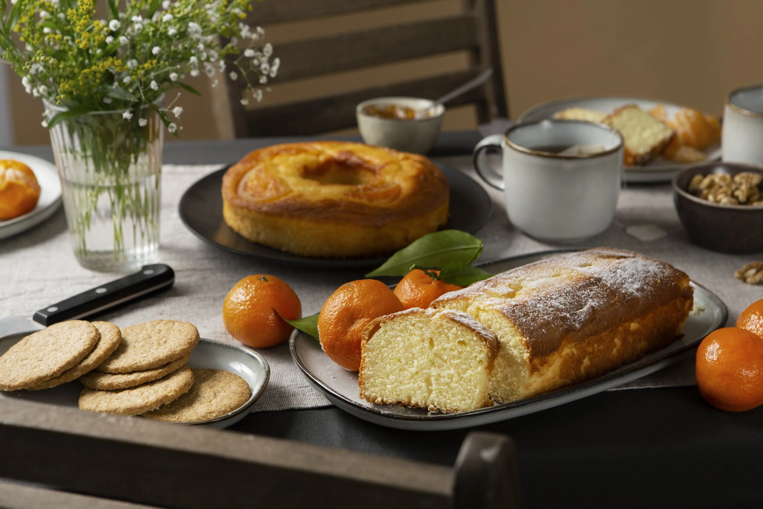 A breakfast table with a loaf of sliced pound cake, fresh tangerines, a cranberry-orange bread, a bundt cake, cookies, a bowl of walnuts, a pot of jam, a cup of coffee, and a vase of white and yellow flowers.