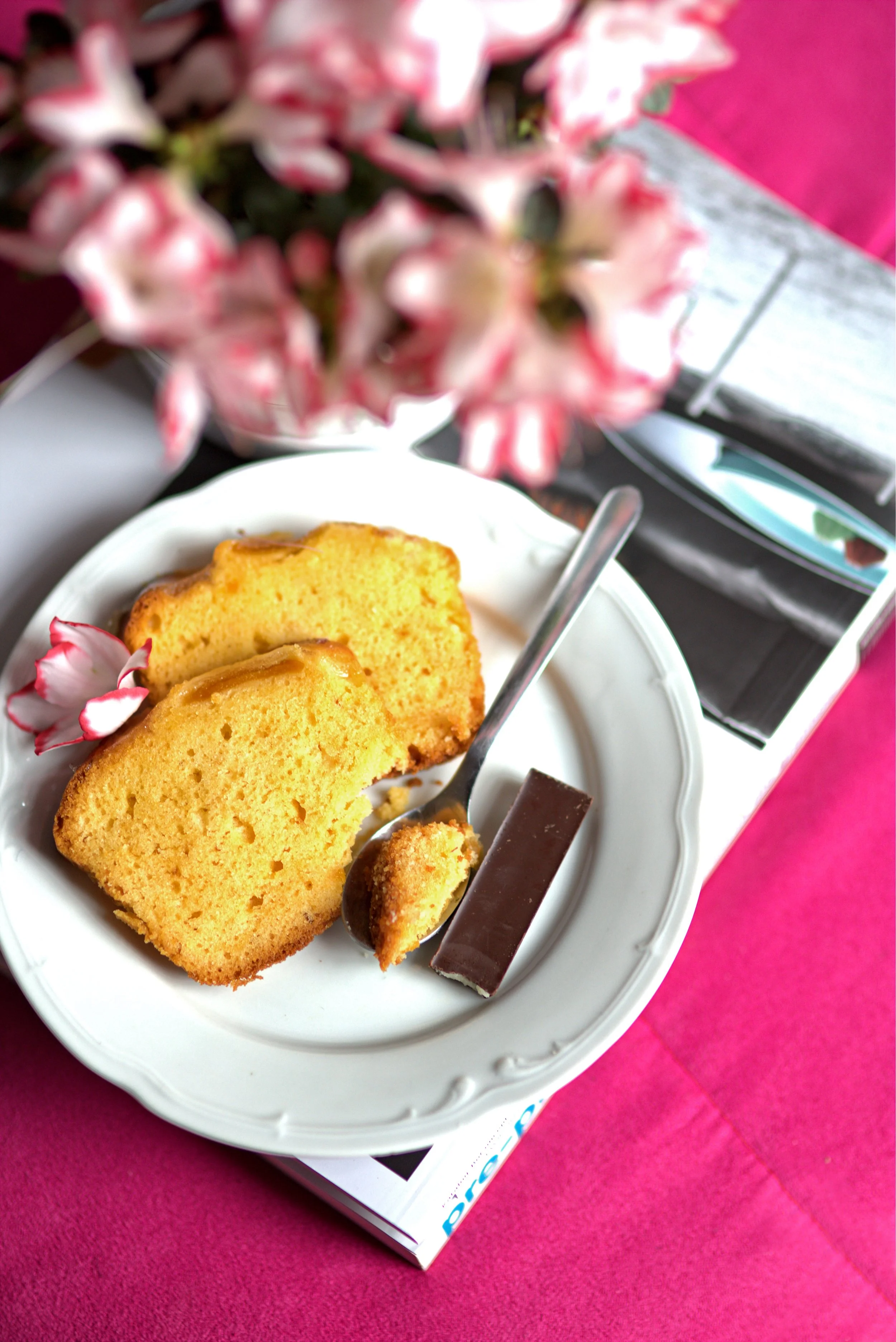 Slices of yellow cake on a white plate with a fork, a chocolate bar, and pink flower petals, set on a pink tablecloth with some flowers and magazines in the background.