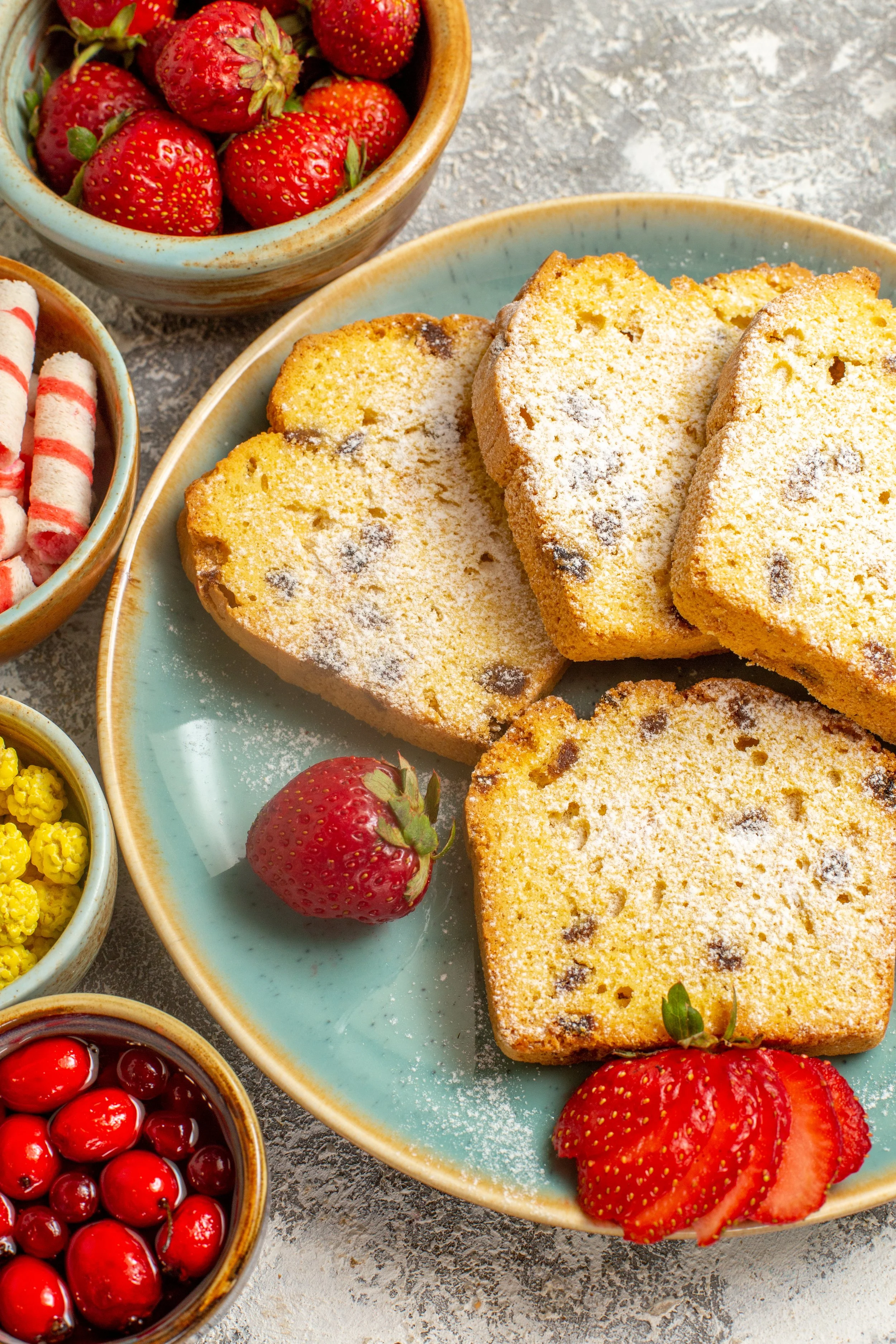 Four slices of lemon poppy seed cake on a turquoise plate, garnished with a whole and sliced strawberry, with bowls of strawberries, red currants, yellow mulberries, and striped marshmallow sticks around.