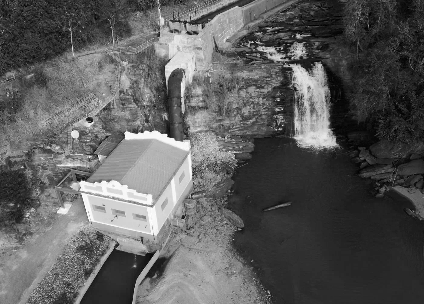 Imagem aérea de uma hidrelétrica com uma casa ao lado de uma represa, água passando por uma comporta e uma cascata ao fundo.