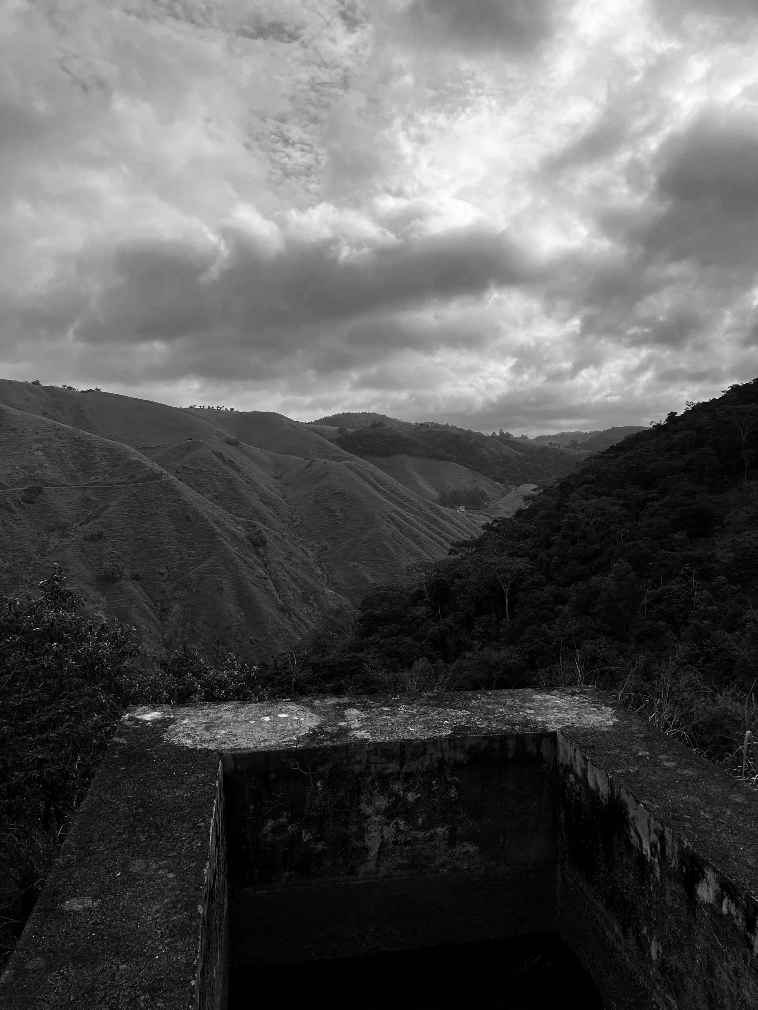 Cenário de montanhas onduladas sob céu nublado visto de uma estrutura de concreto na frente.