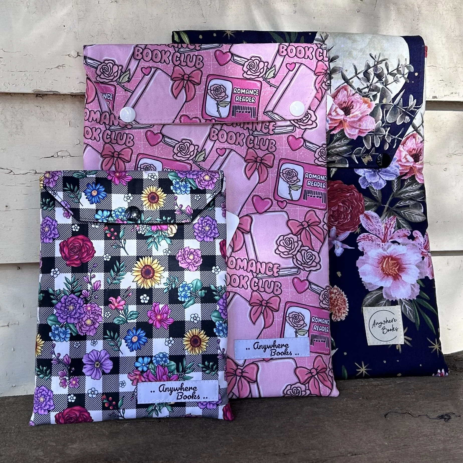 Three decorative fabric book covers with floral and themed designs, displayed against a white wooden background.