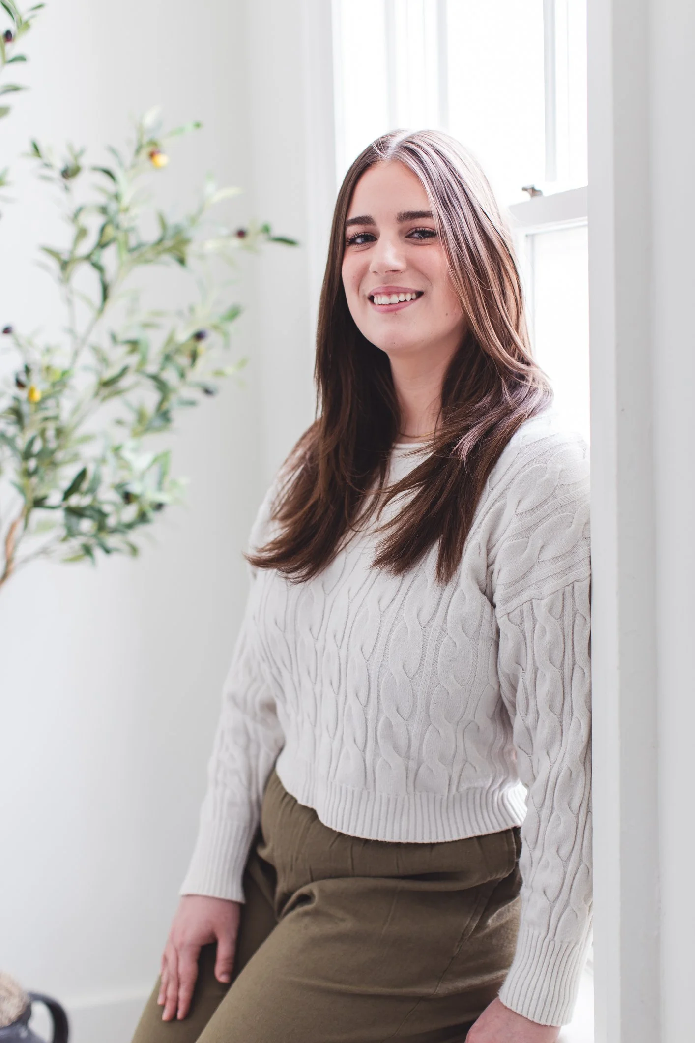 A young woman with long brown hair smiling and sitting by a window, wearing a white cable-knit sweater and brown pants.
