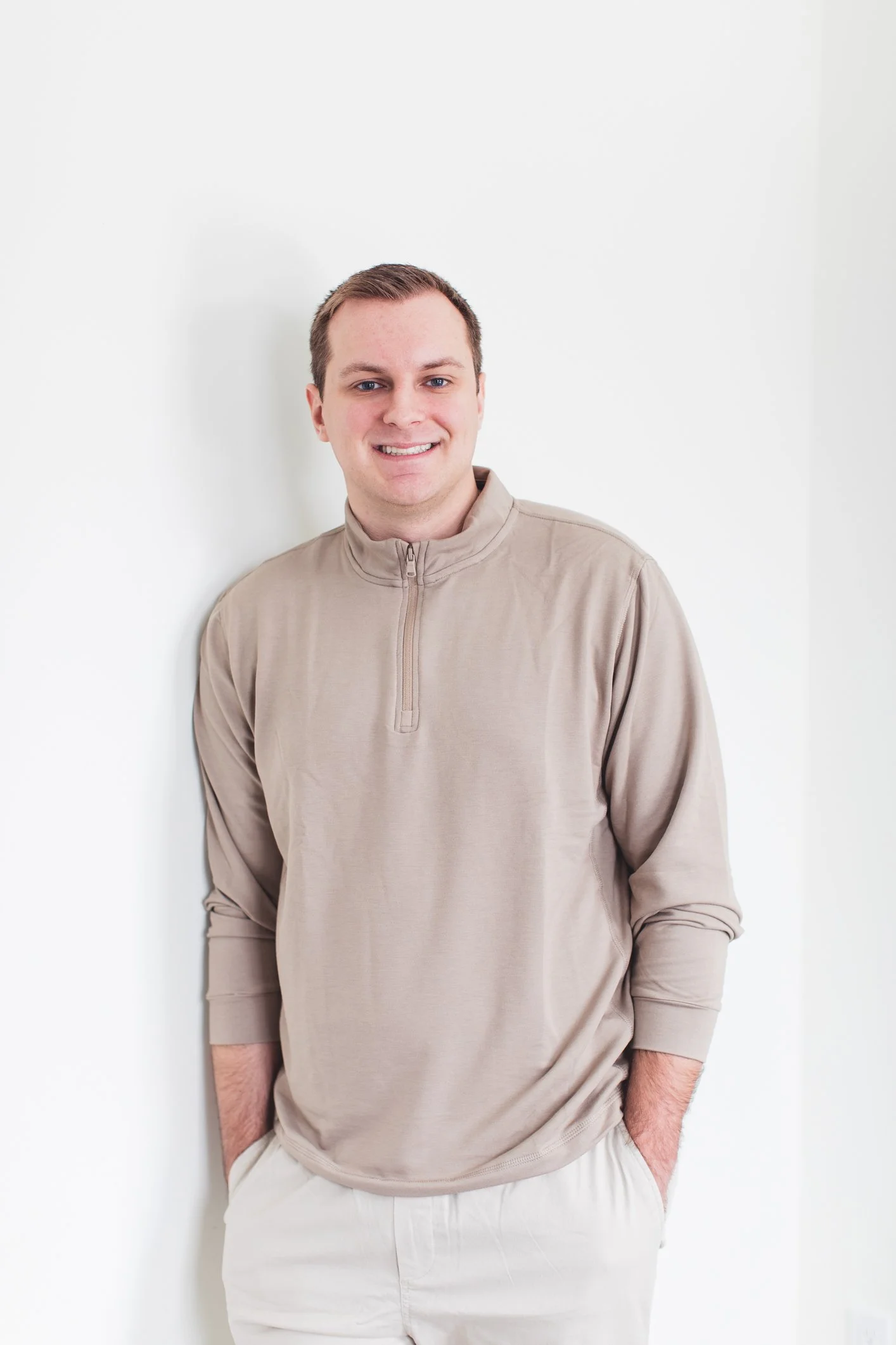 Portrait of a smiling man with short brown hair, wearing a beige quarter zipper pullover and light-colored pants, standing against a plain white wall.