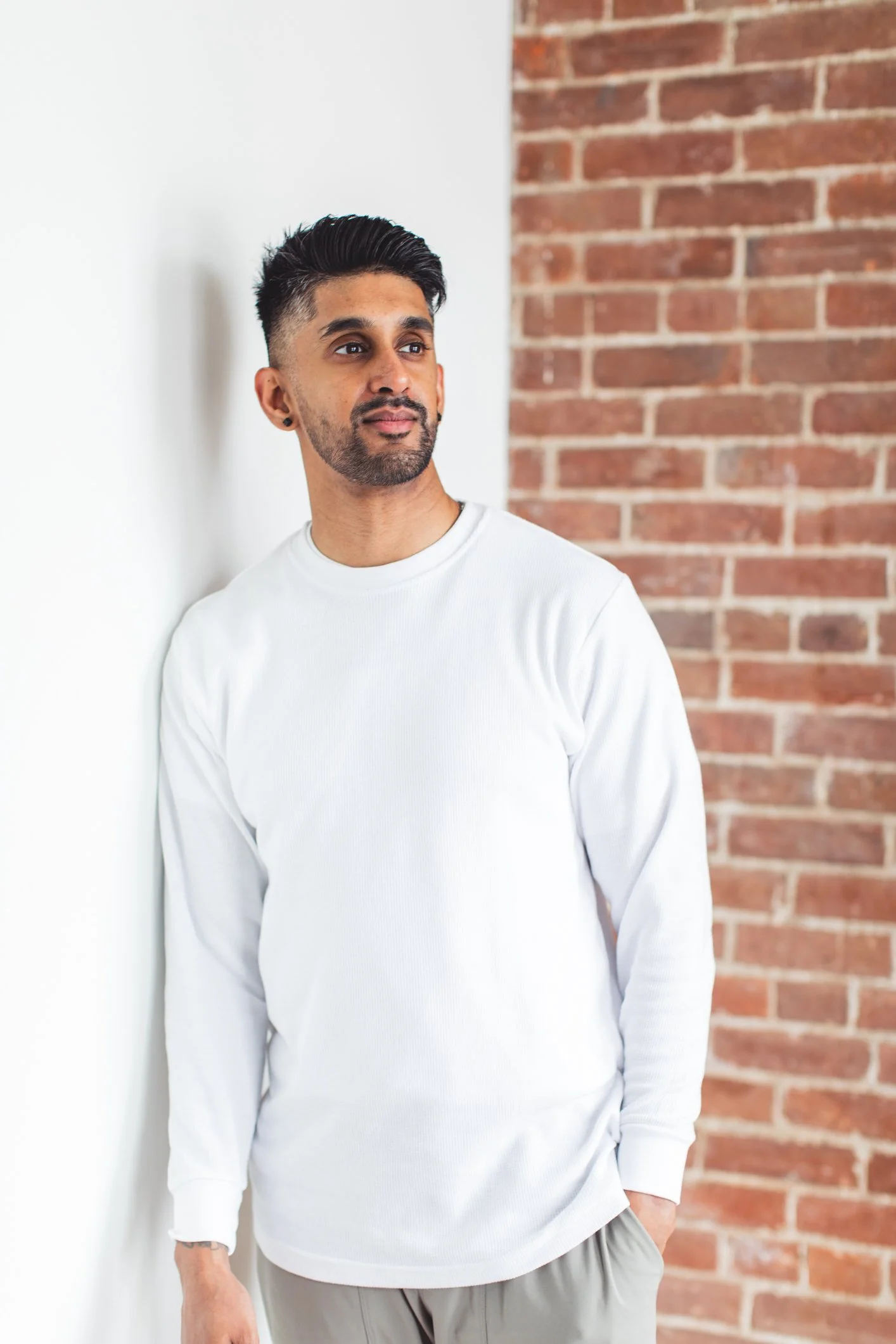 A man with dark hair and beard, wearing a white long-sleeve shirt and light-colored pants, standing against a white wall and red brick wall, looking to his left.