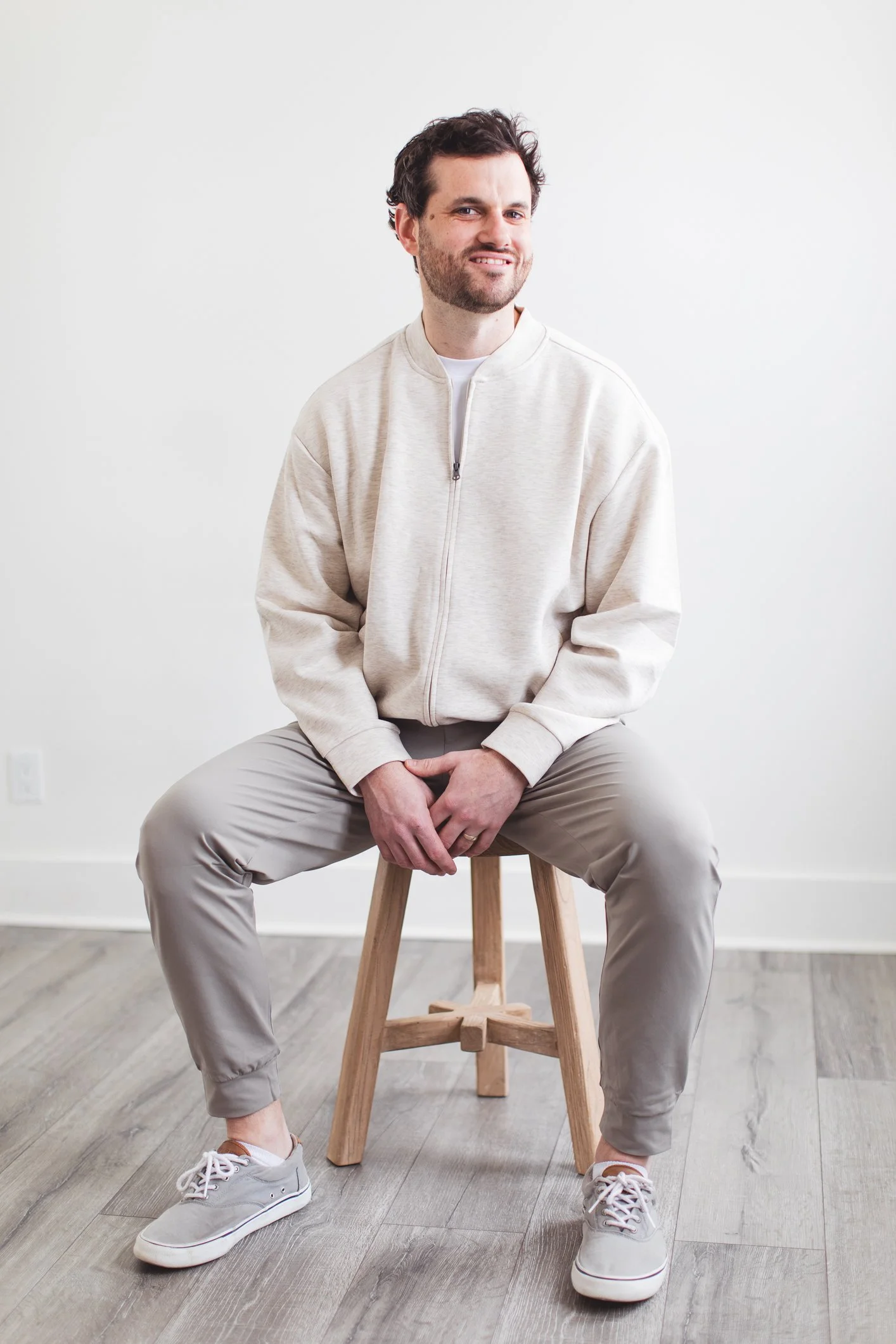A man sitting on a wooden stool against a white wall, wearing a light beige zip-up sweatshirt, beige pants, and light gray sneakers, smiling and looking at the camera.
