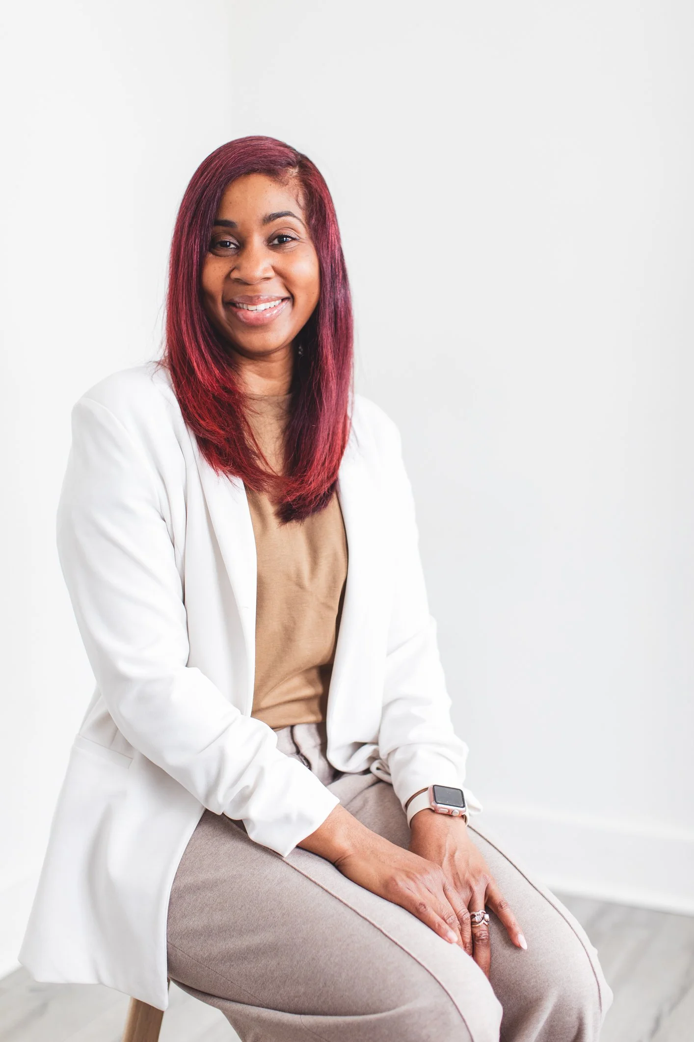 A woman with red hair smiling, wearing a white blazer, sitting on a beige chair with a smartwatch on her left wrist, against a plain white background.