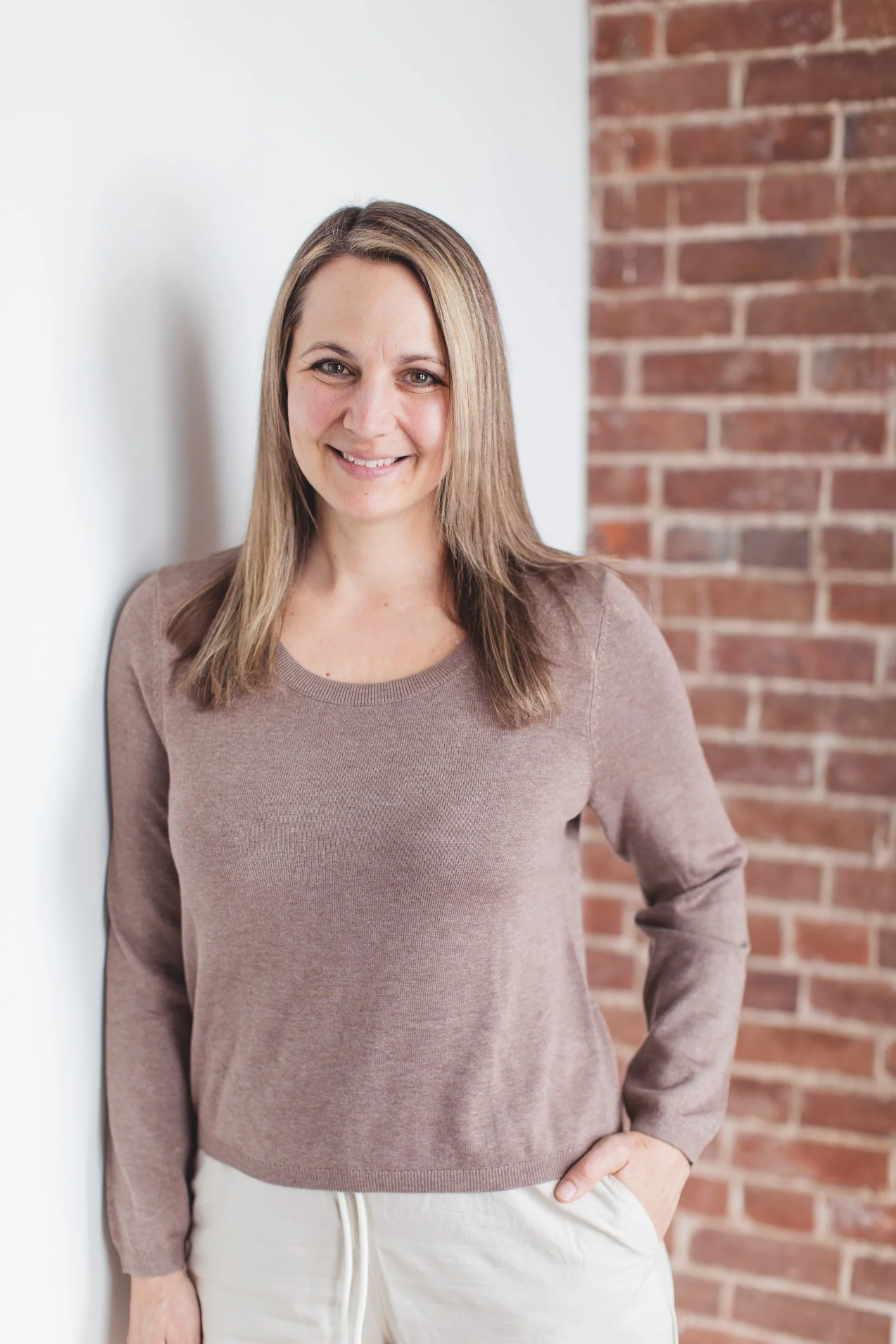 A woman with light brown hair, smiling, standing against a white wall and a brick wall, wearing a beige sweatshirt and beige pants.