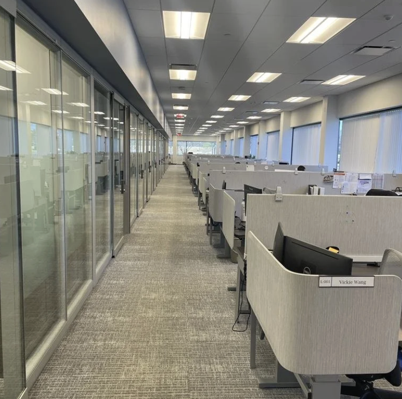 Empty office workstations with desks and chairs in a modern office space, glass-walled meeting rooms on the left, and large windows on the right.