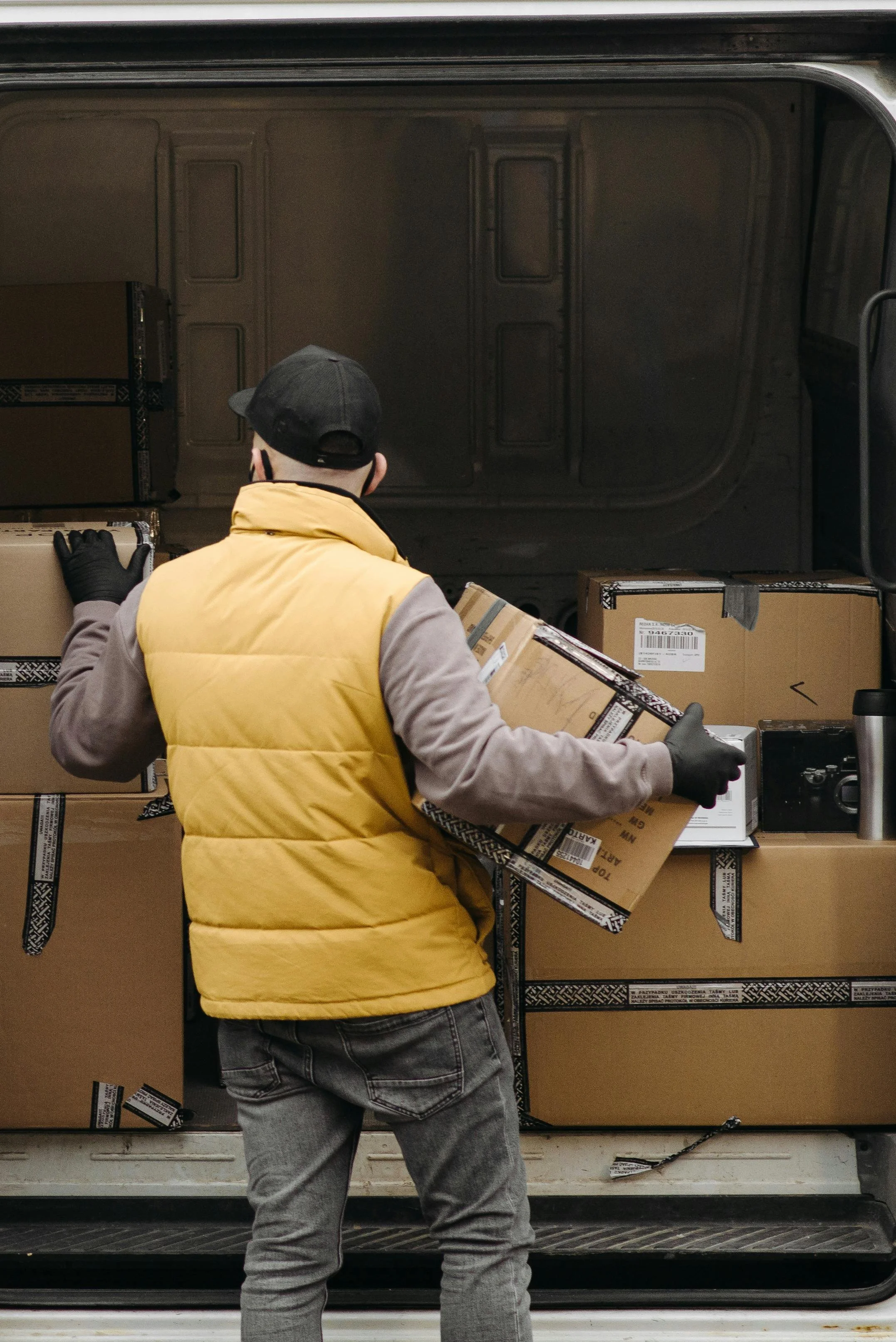 A delivery worker loading boxes into a truck, wearing a yellow vest, gloves, and a black cap.