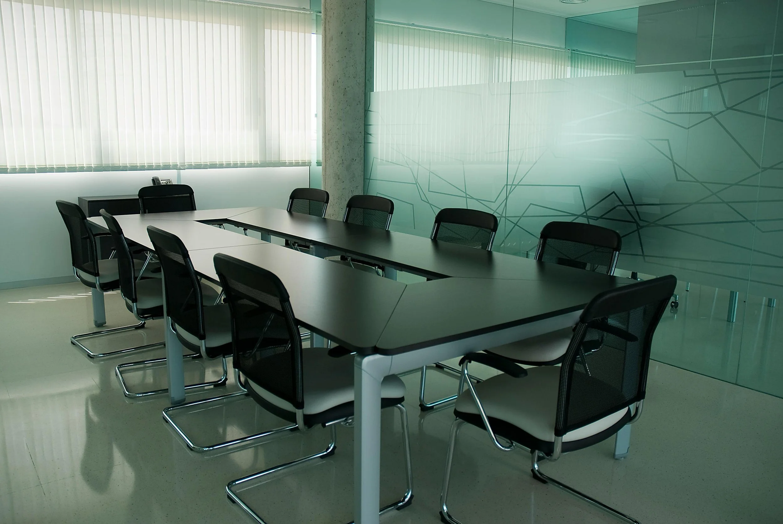 Empty modern conference room with a long black table surrounded by ten black office chairs, large window with vertical blinds, frosted glass wall with geometric pattern.